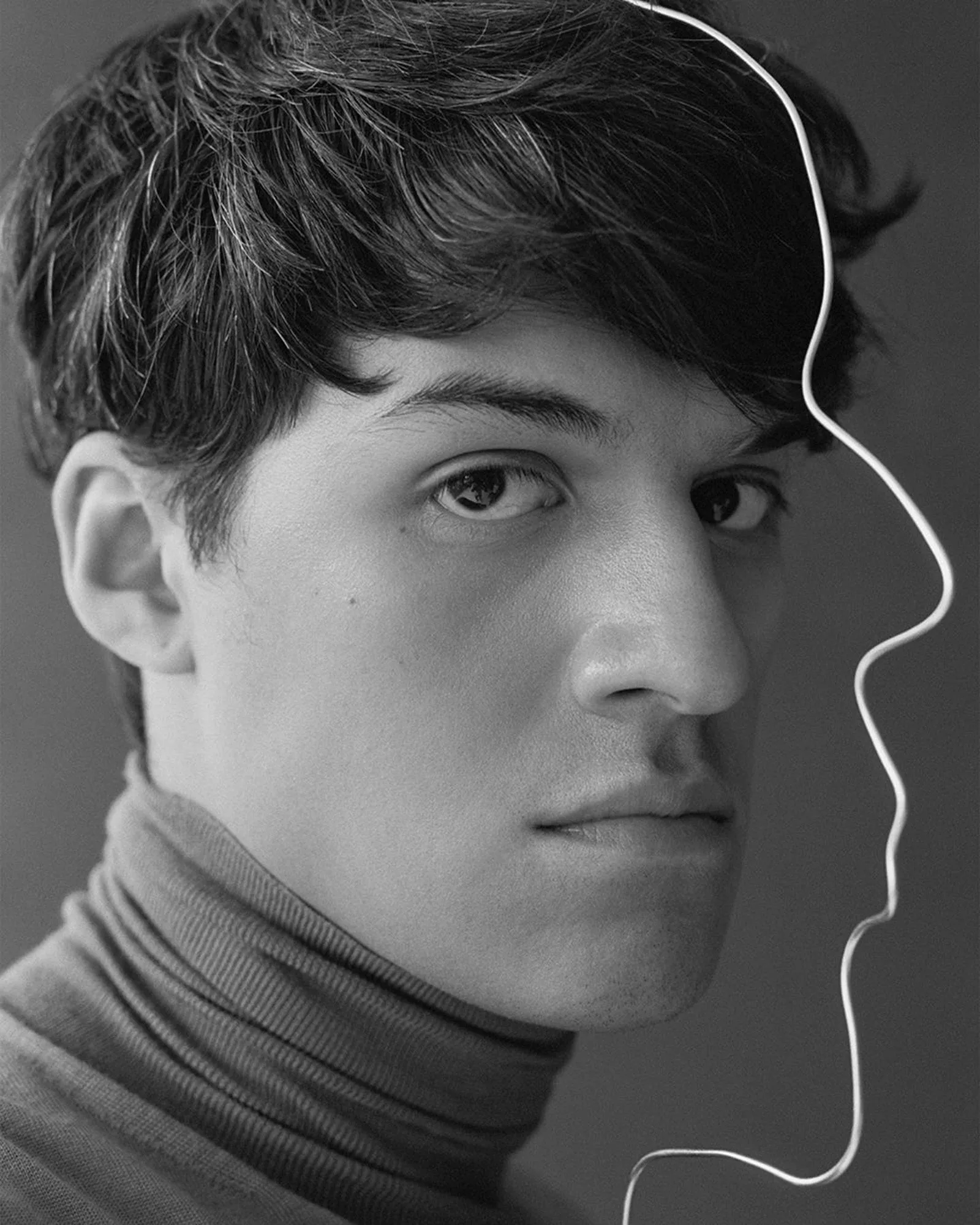 Black and white portrait of a man in a turtleneck beside a wire sculpture tracing a face profile, for The Last Magazine.