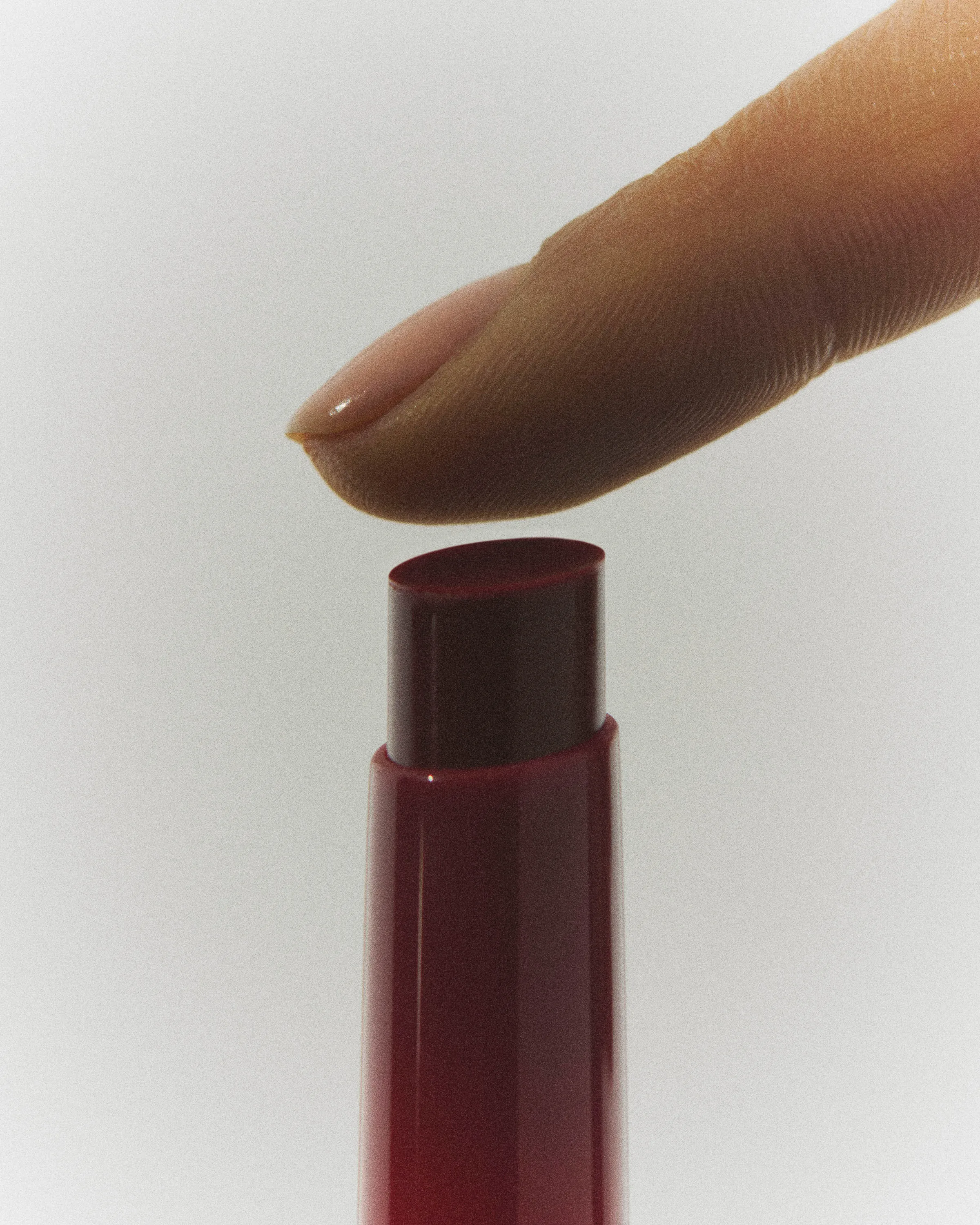 Fingertip hovering over a berry-red lip stick bullet in a glossy red tube on a pale grey backdrop, for Kylie Cosmetics.