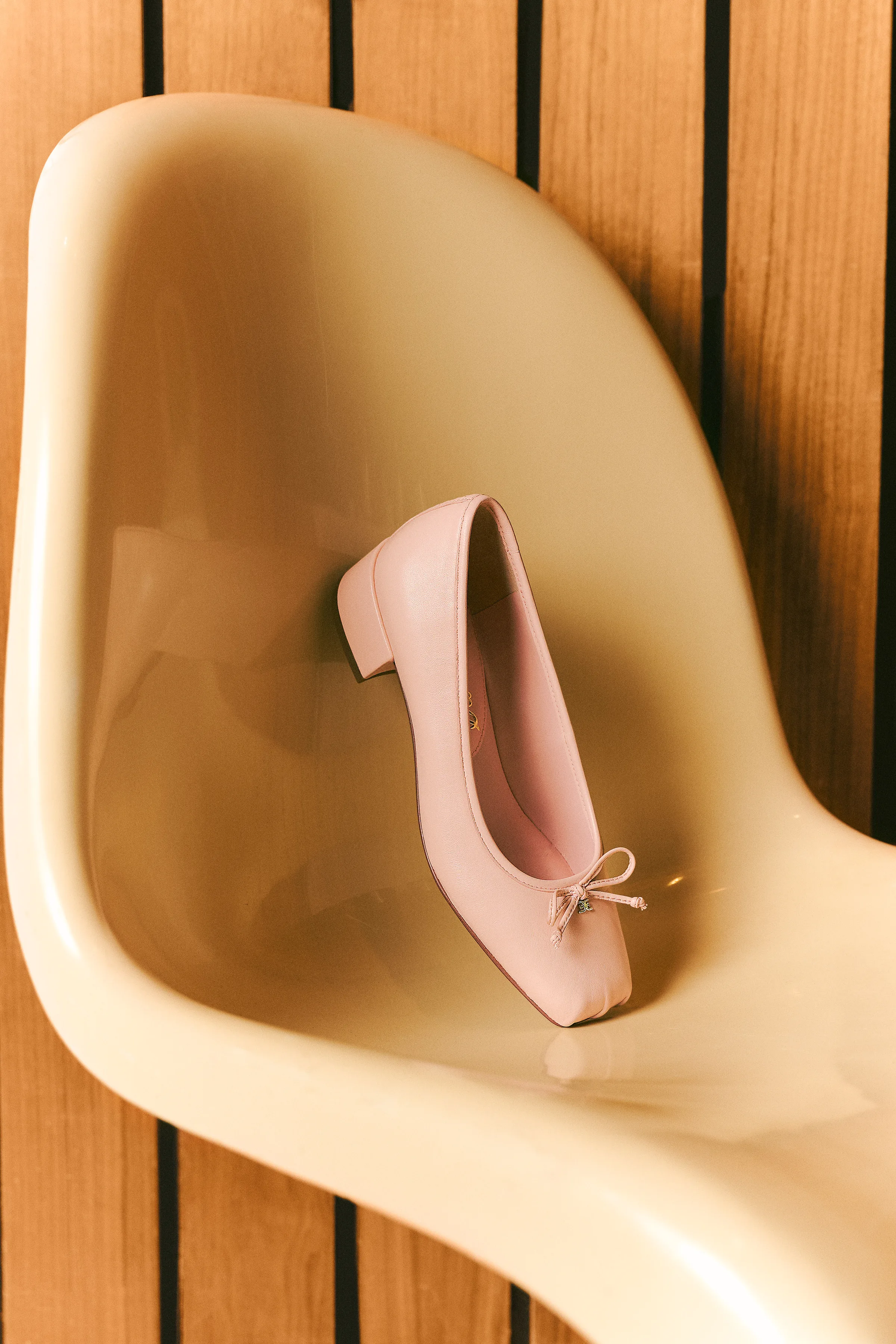 Pink leather ballet flat with a bow and low heel resting on a cream molded chair against wood slats, for Sam Edelman.