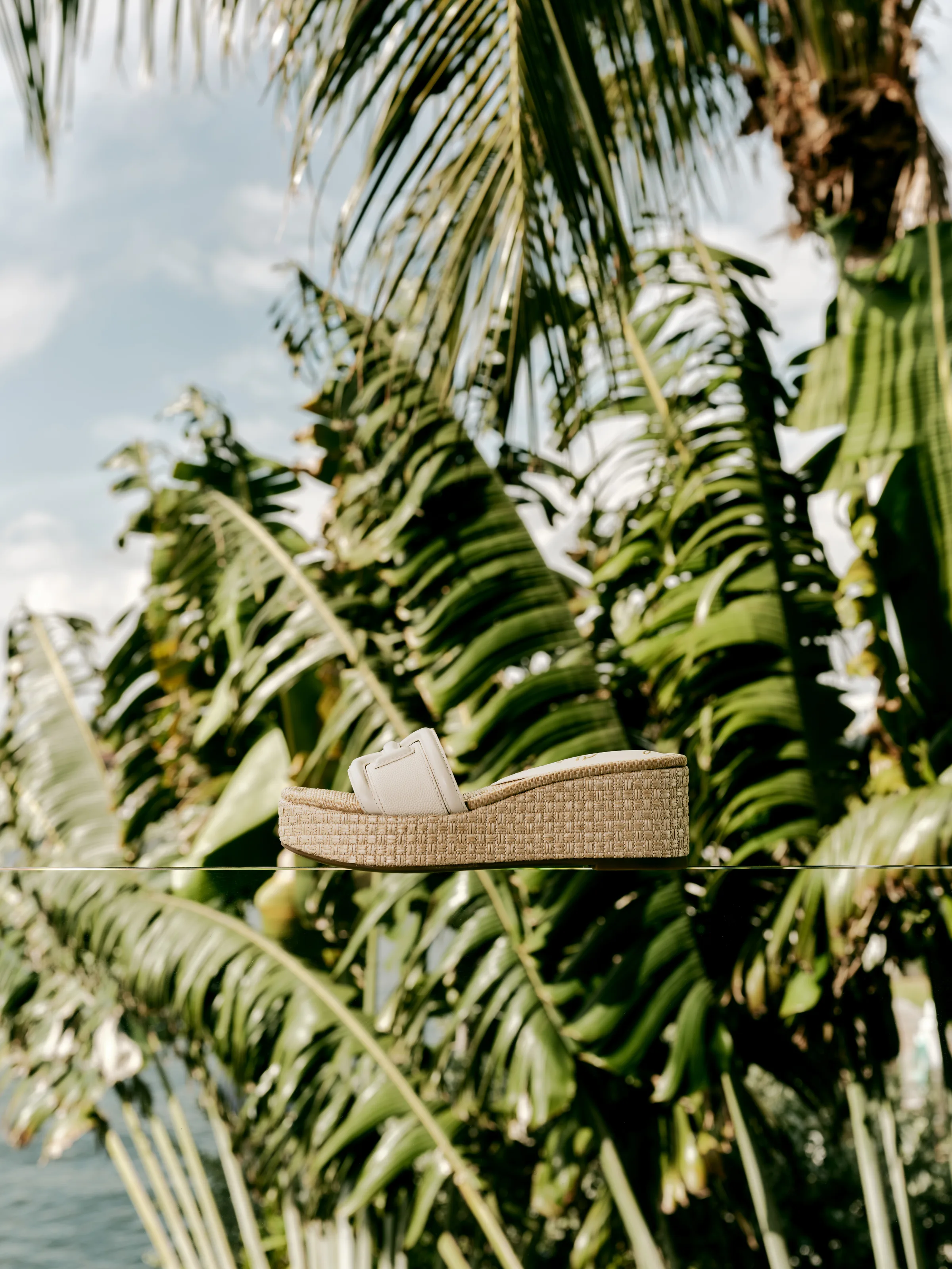 White strap platform slide with woven raffia wedge balanced on a glass ledge before palm trees and sky, for Sam Edelman.