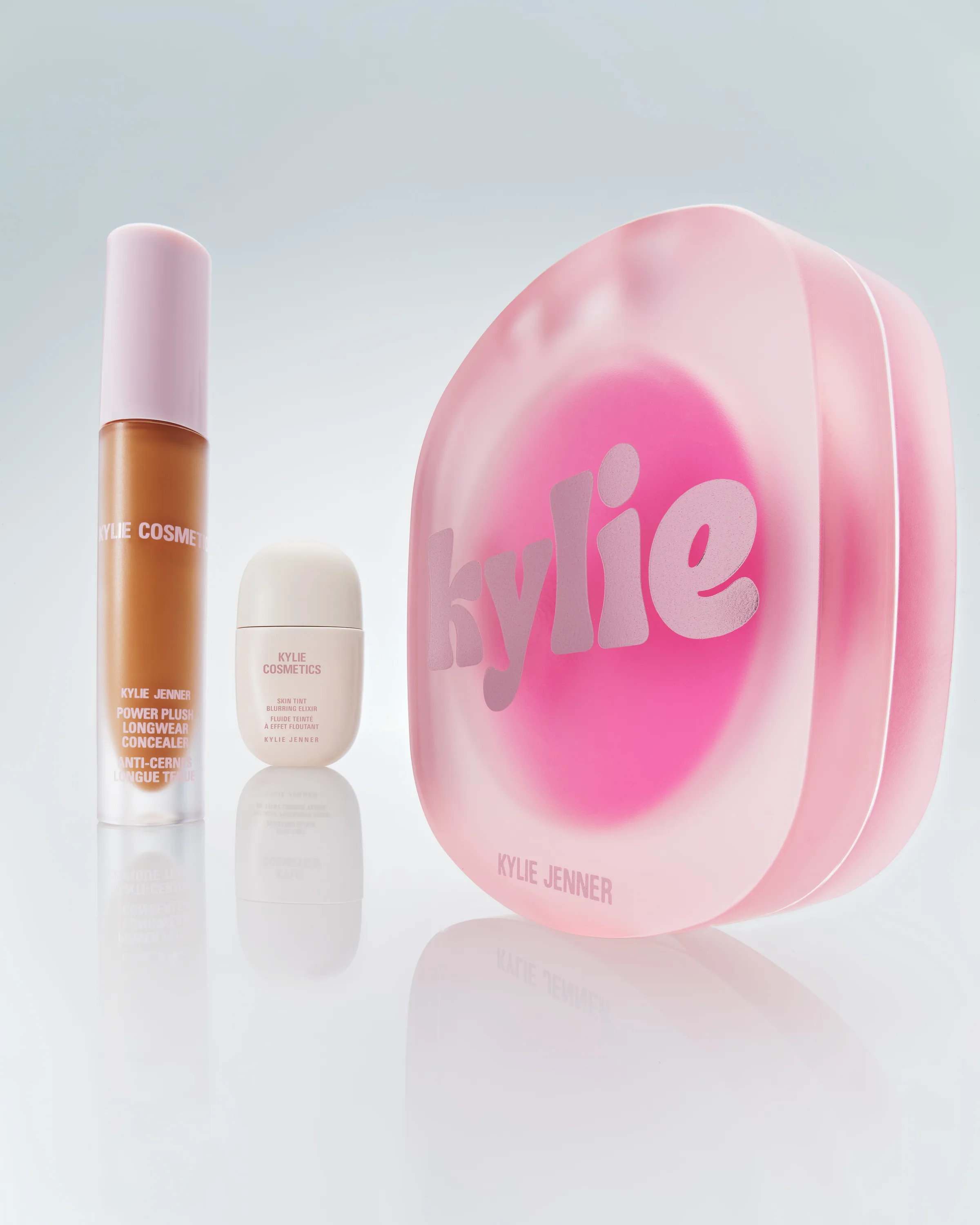 Concealer, skin tint bottle and frosted pink glow balm compact on a glossy pale surface with reflections, for Kylie Cosmetics.