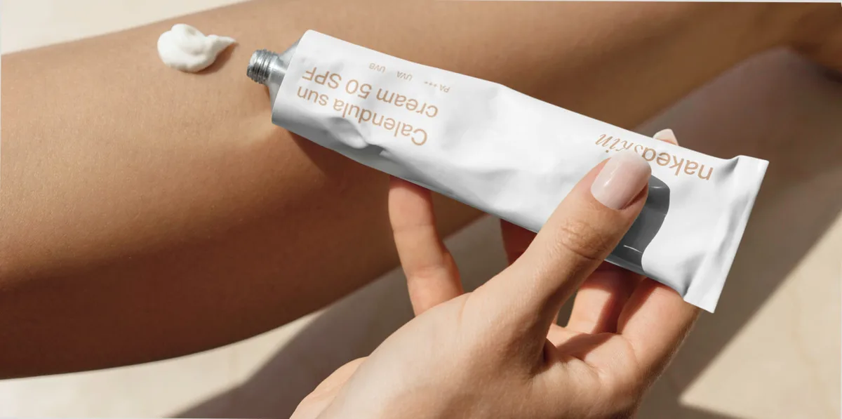Hand holding a white Calendula SPF 50 sun cream tube beside a dab of cream on a tanned leg, for Naked Skin.