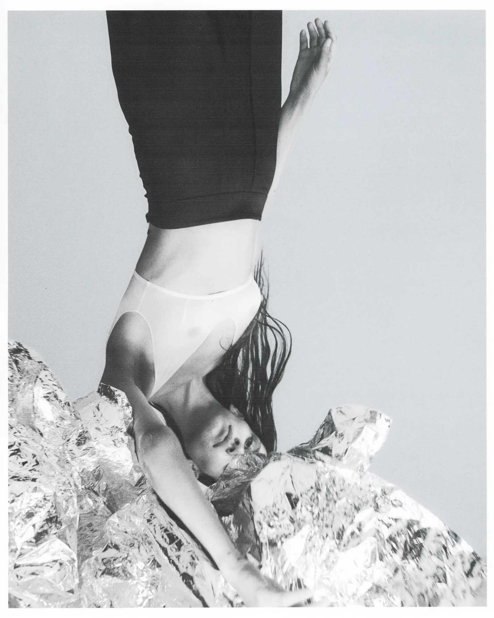Black-and-white editorial of a model posed upside down in a sheer bodysuit over crumpled silver foil, for The Last Magazine.