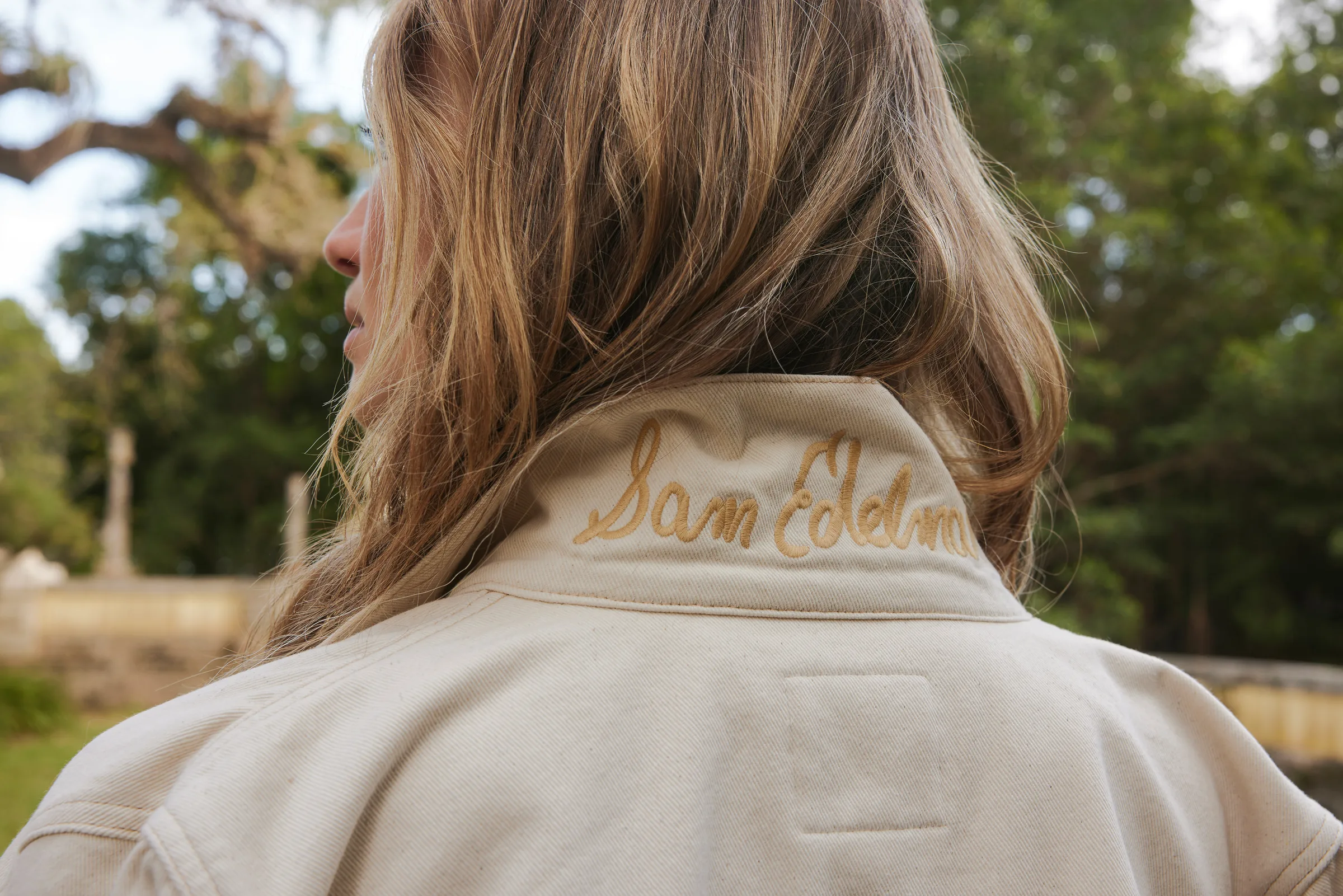 Woman seen from behind in a cream canvas jacket with gold script embroidered on the collar, outdoors among trees, for Sam Edelman.