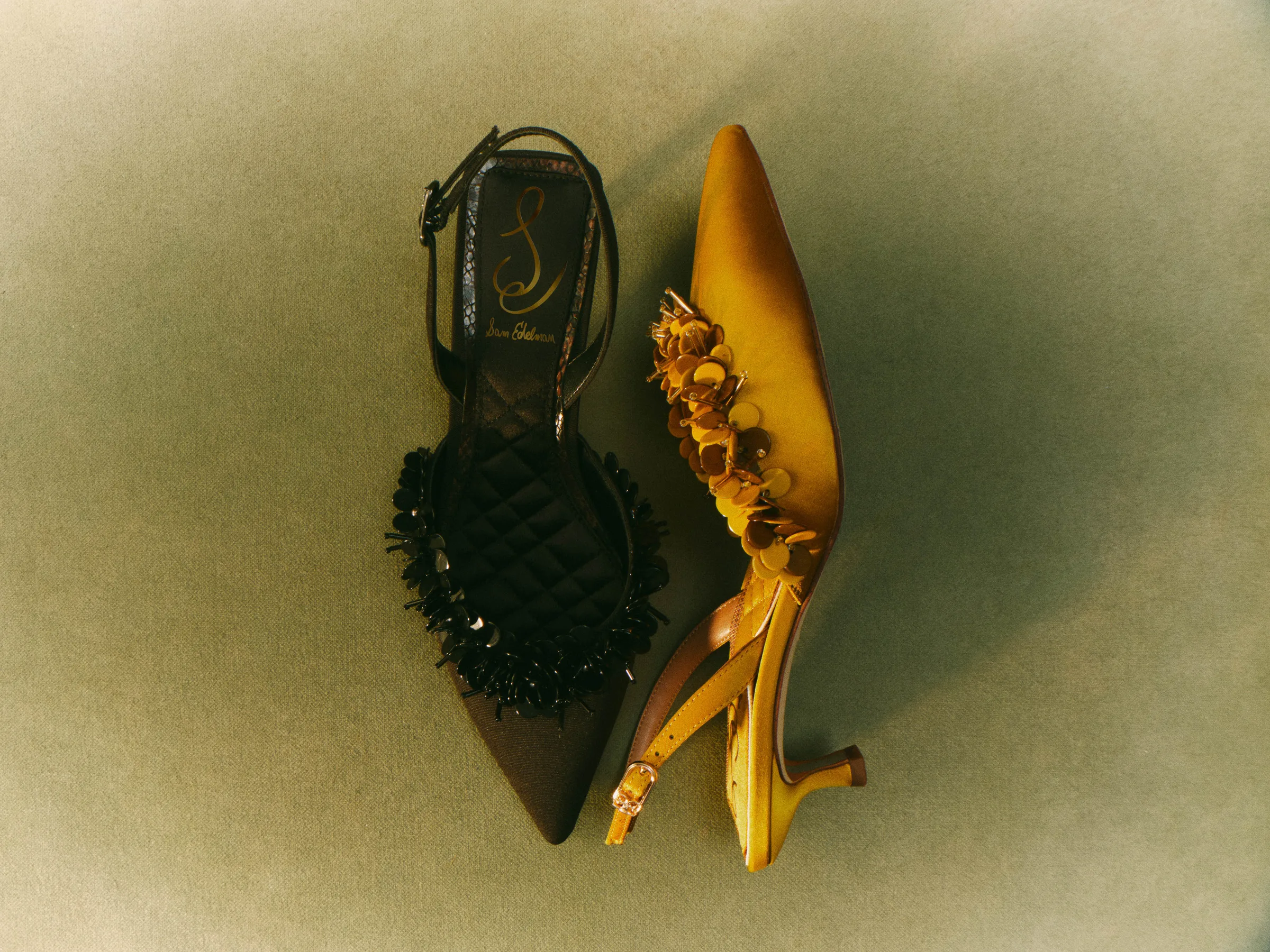 Black beaded and yellow sequin-trimmed kitten heel slingbacks laid on a muted green backdrop, for Sam Edelman.