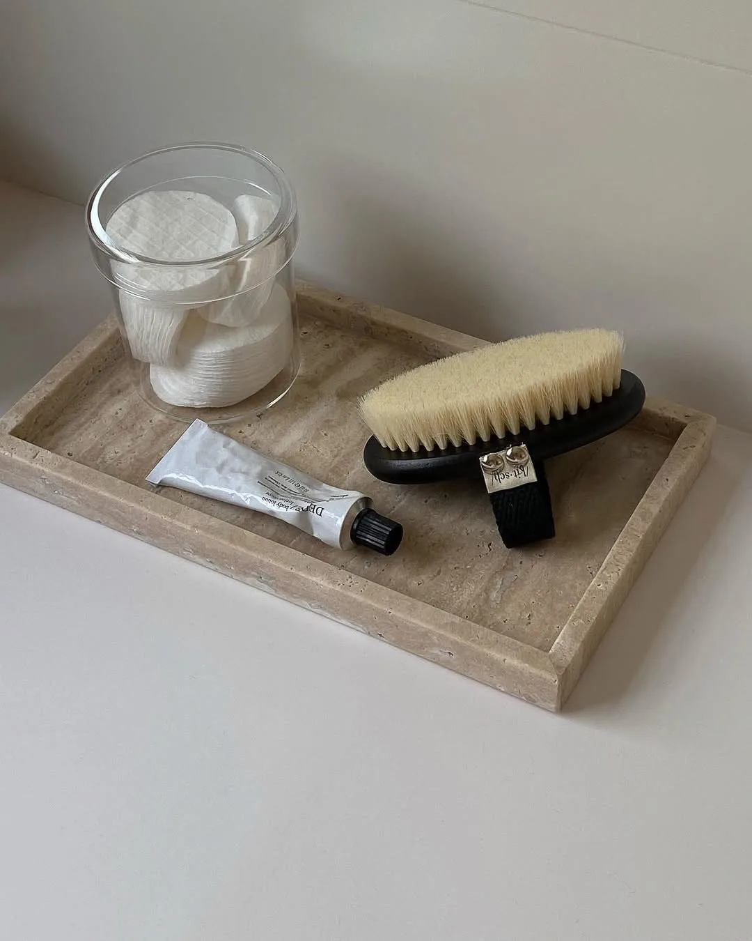 Travertine tray holding a dry body brush, a tube of hand cream and a glass jar of cotton pads on a white counter.