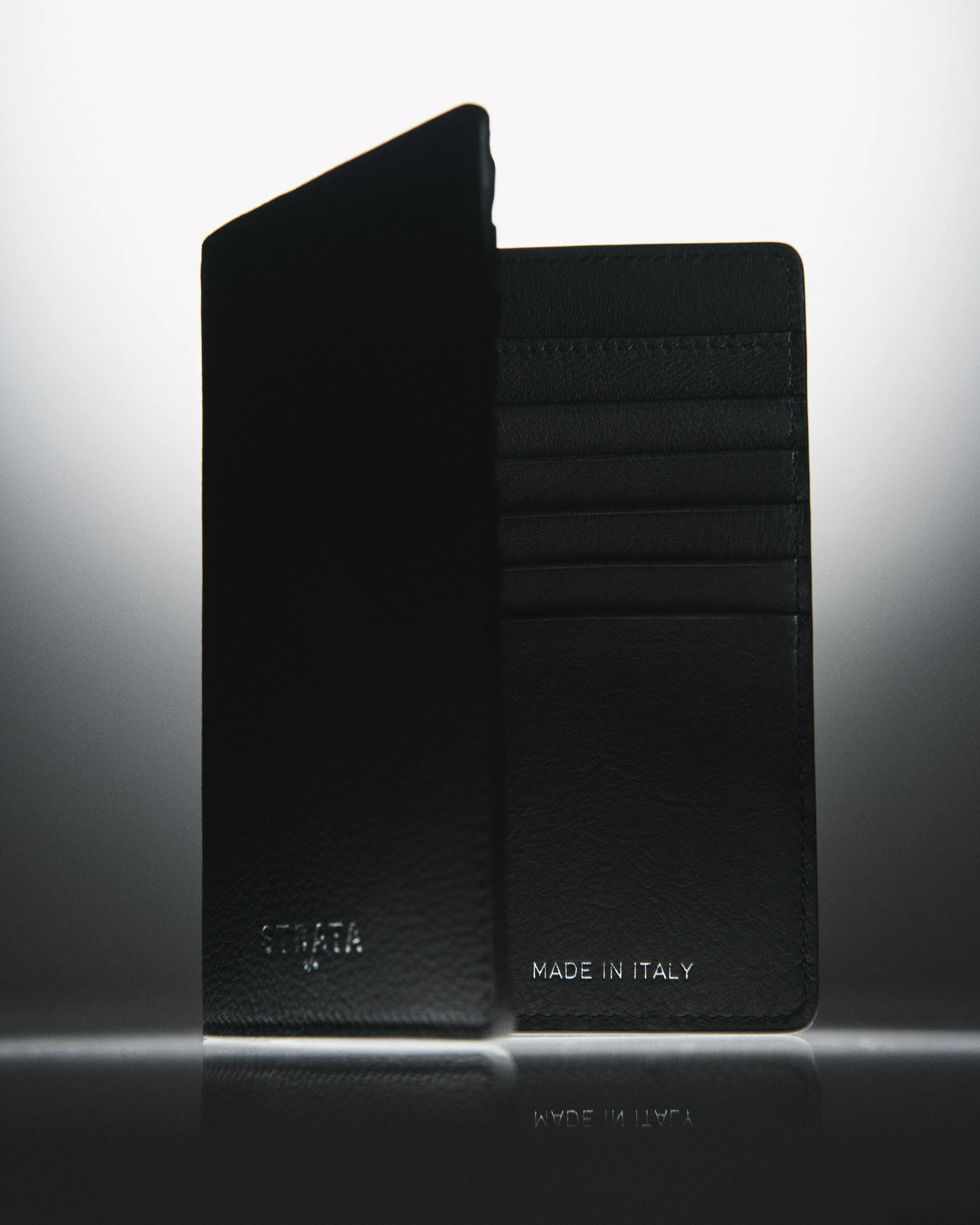 Open black leather travel sleeve with card slots, standing on a reflective surface against a soft grey glow, for Strata.