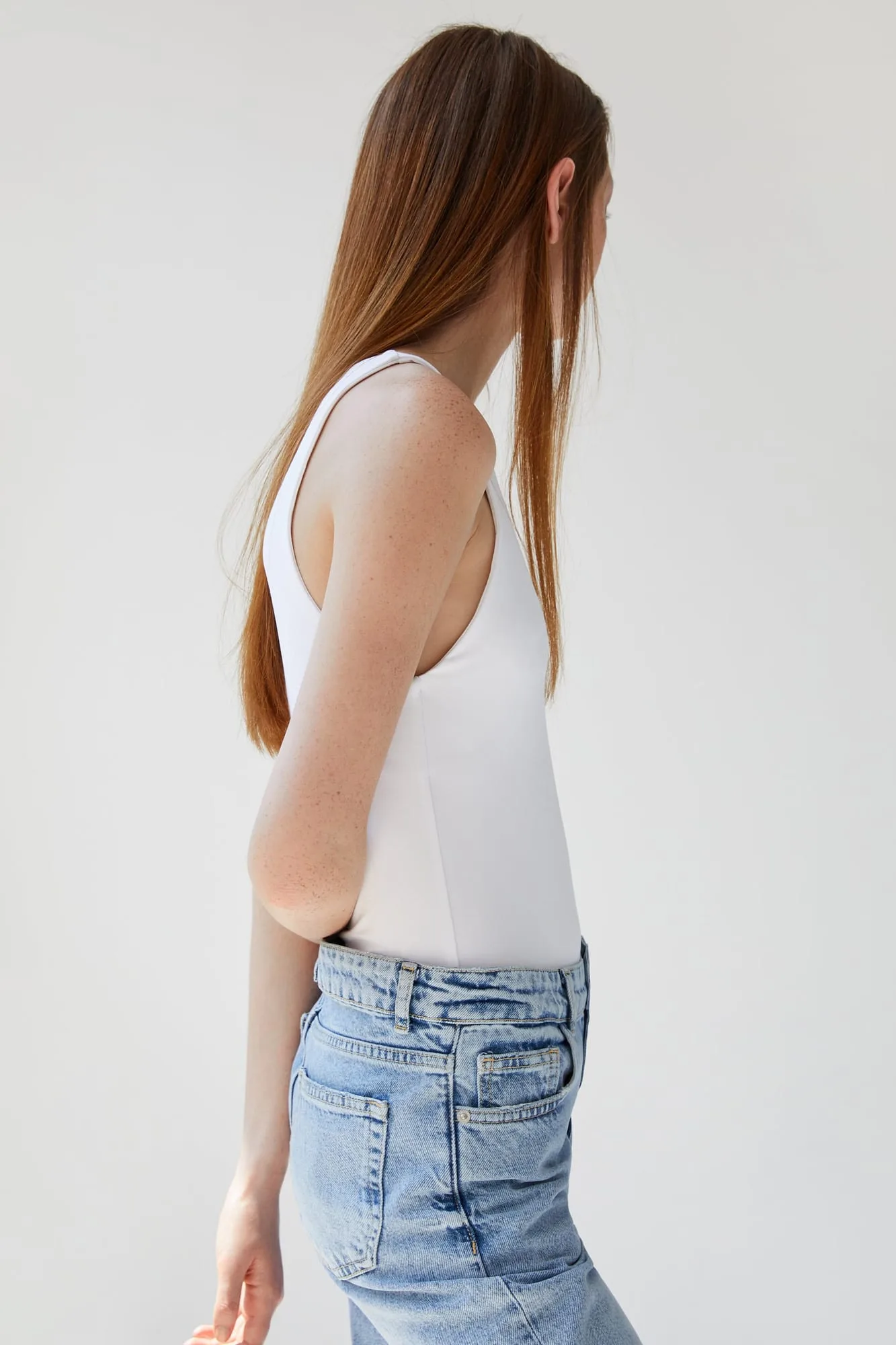 Model in profile wearing a white sleeveless bodysuit and light-wash jeans against a plain grey backdrop, for Zara.