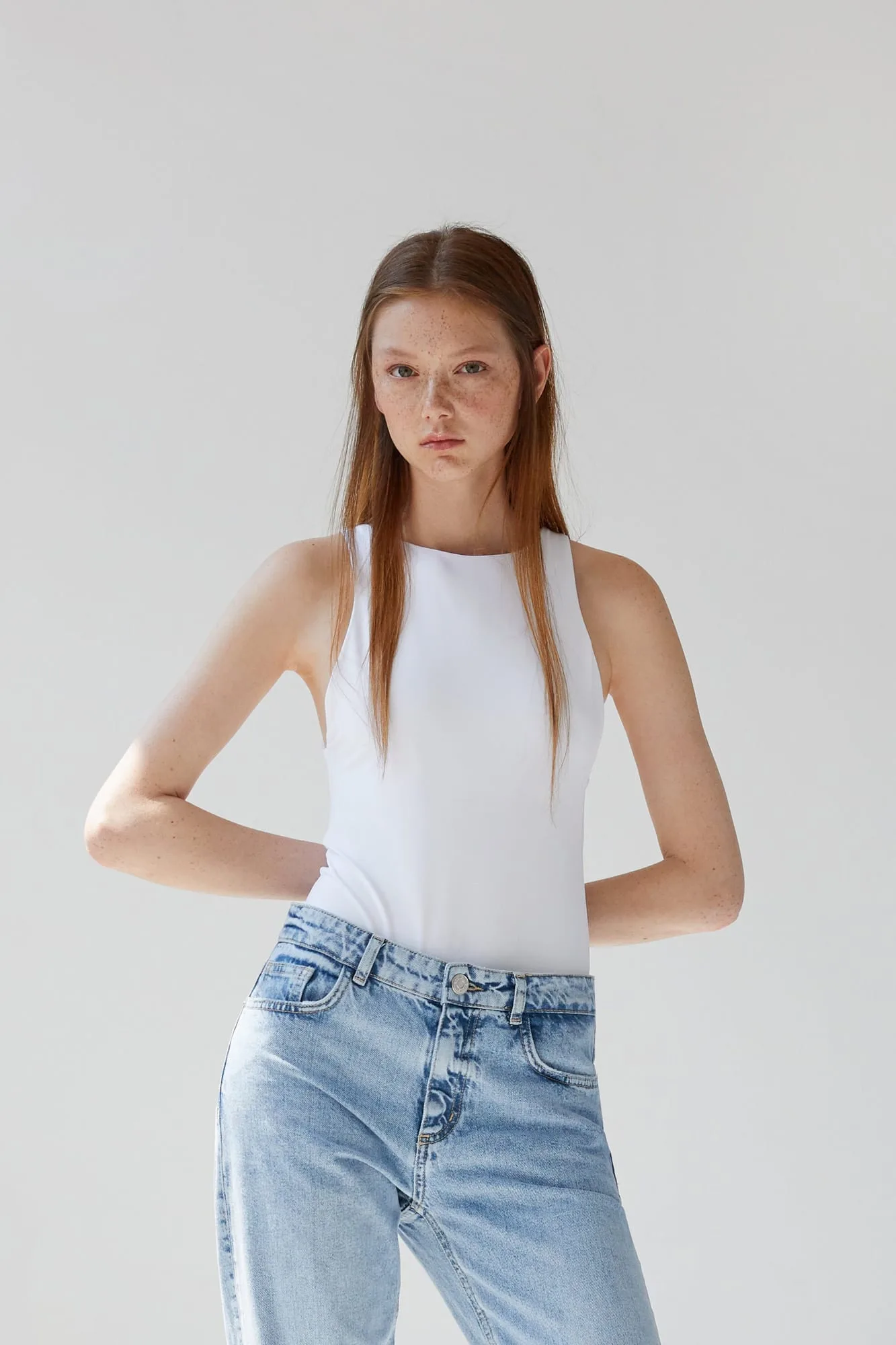 Red-haired model in a white sleeveless top and light-wash jeans, hands behind her back, on a plain grey backdrop, for Zara.