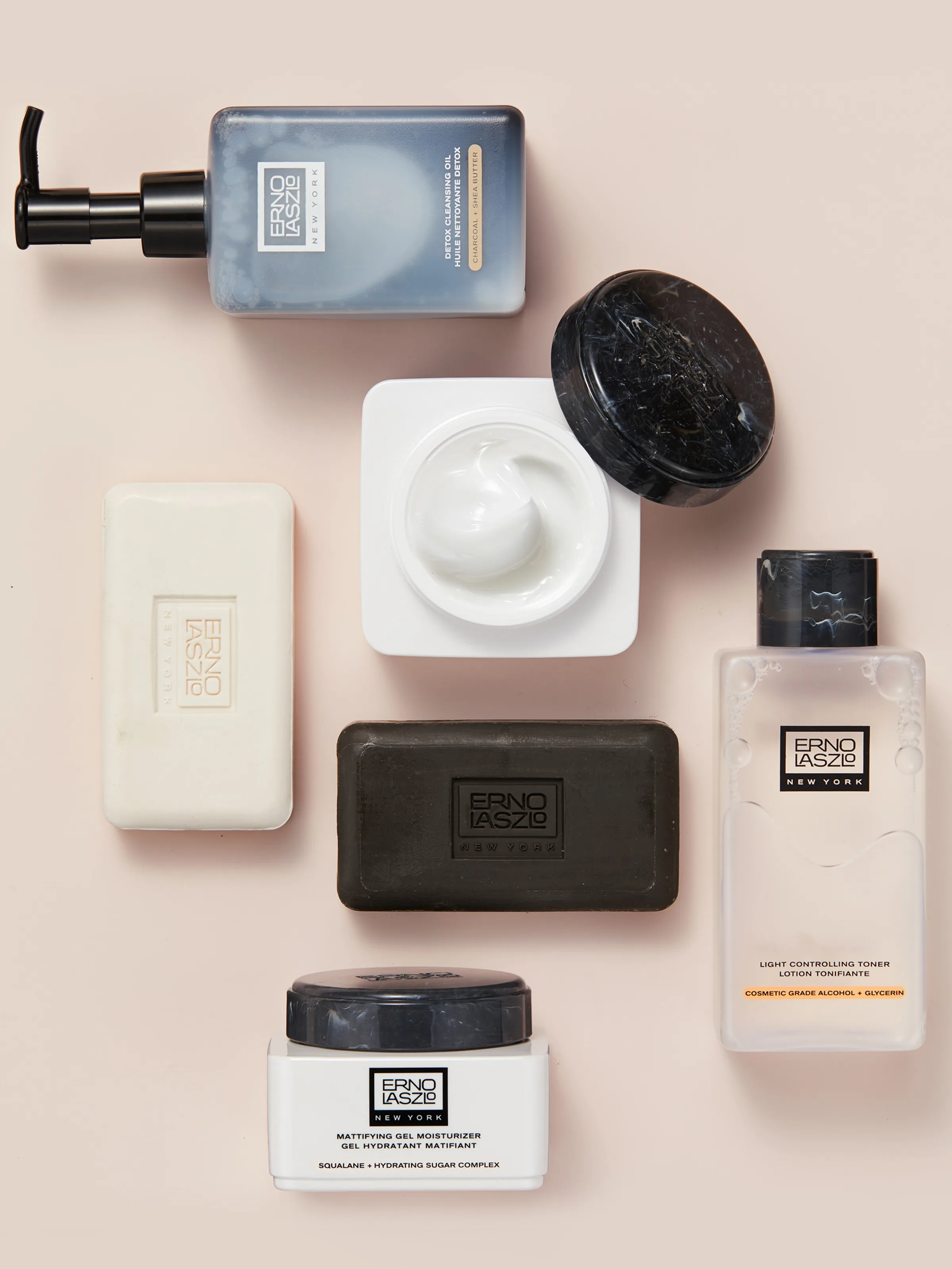Flat lay of cleansing oil, cream jar, black and white soap bars, toner and gel moisturizer on blush pink, for Erno Laszlo.