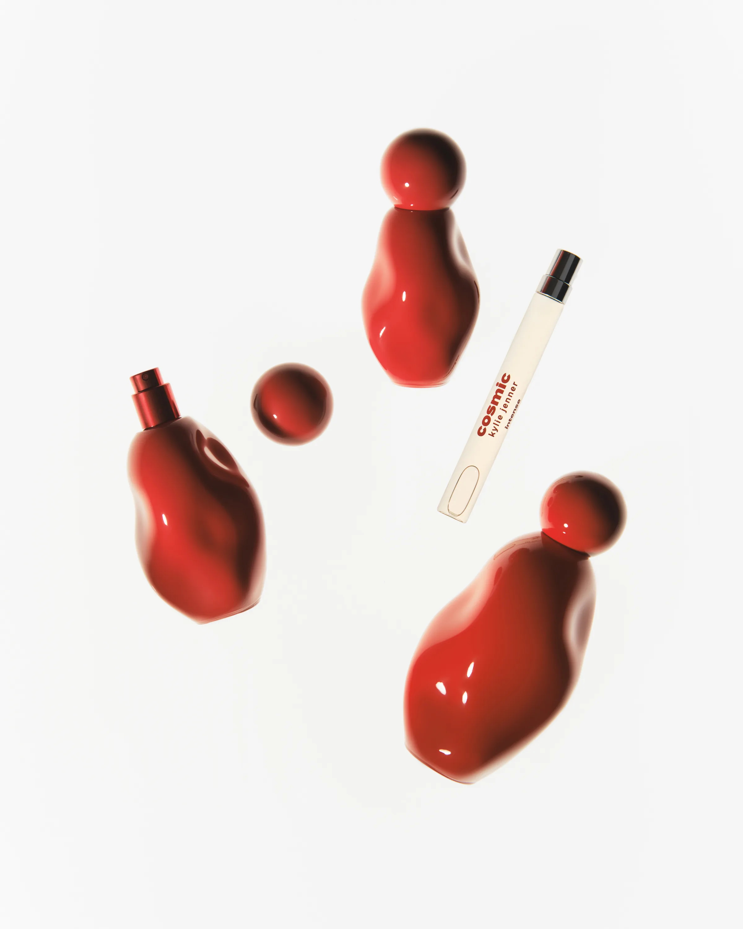 Glossy red sculpted Cosmic perfume bottles, a loose cap and a travel spray arranged on white, for Kylie Cosmetics.