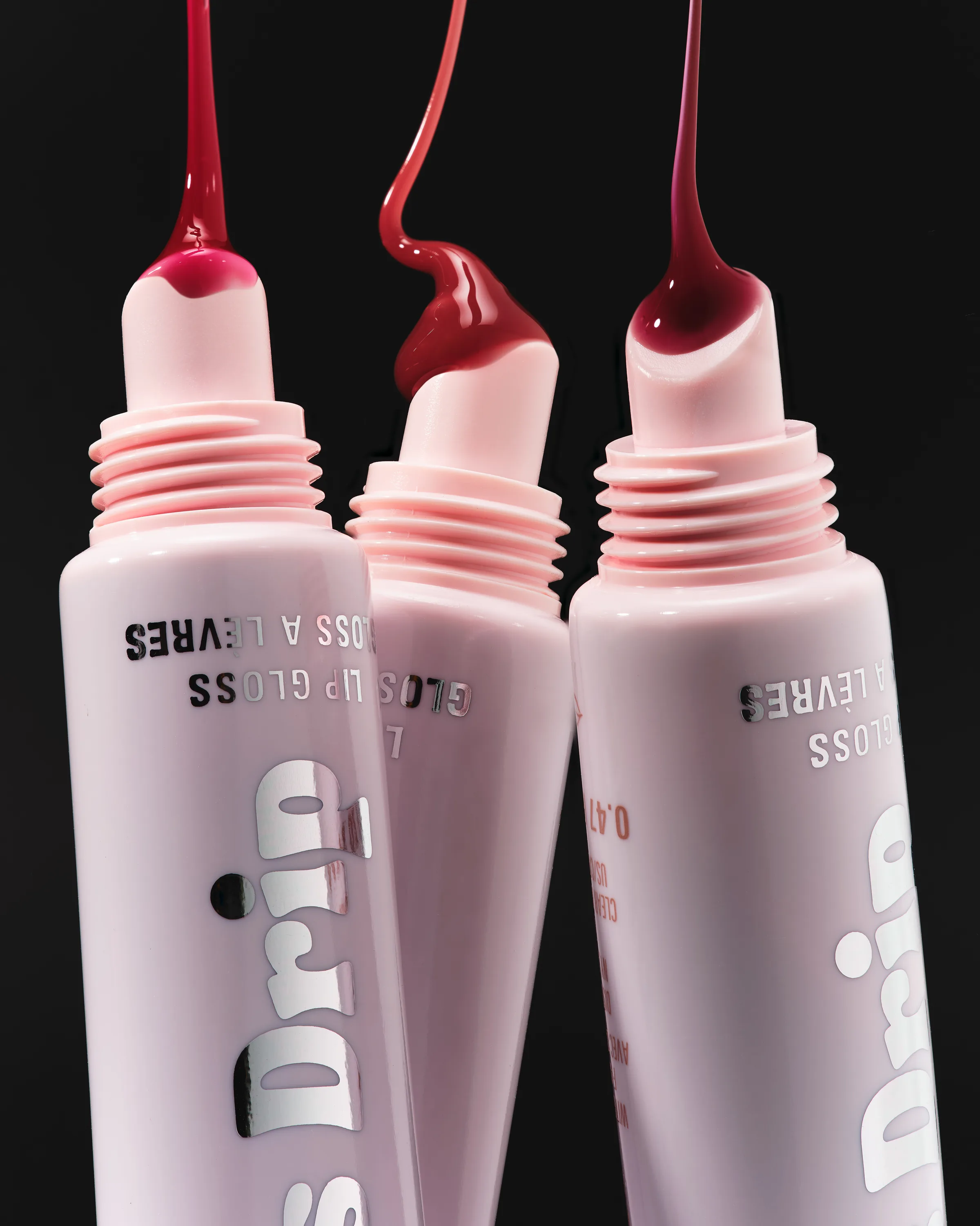 Three pink Gloss Drip lip gloss tubes with red gloss dripping from the tips against a black background, for Kylie Cosmetics.