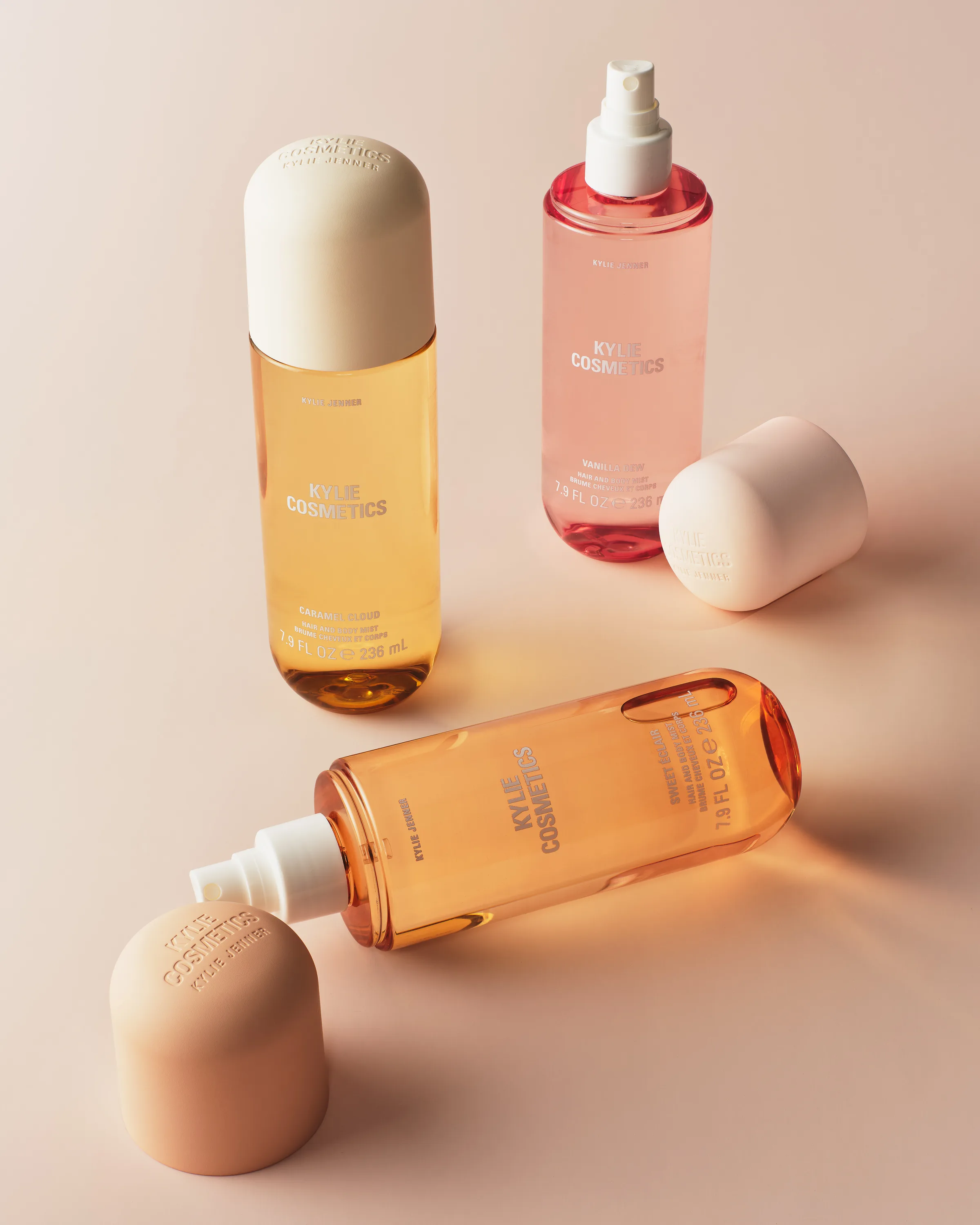 Three hair and body mist bottles in caramel, pink and peach with caps off on a blush background, for Kylie Cosmetics.