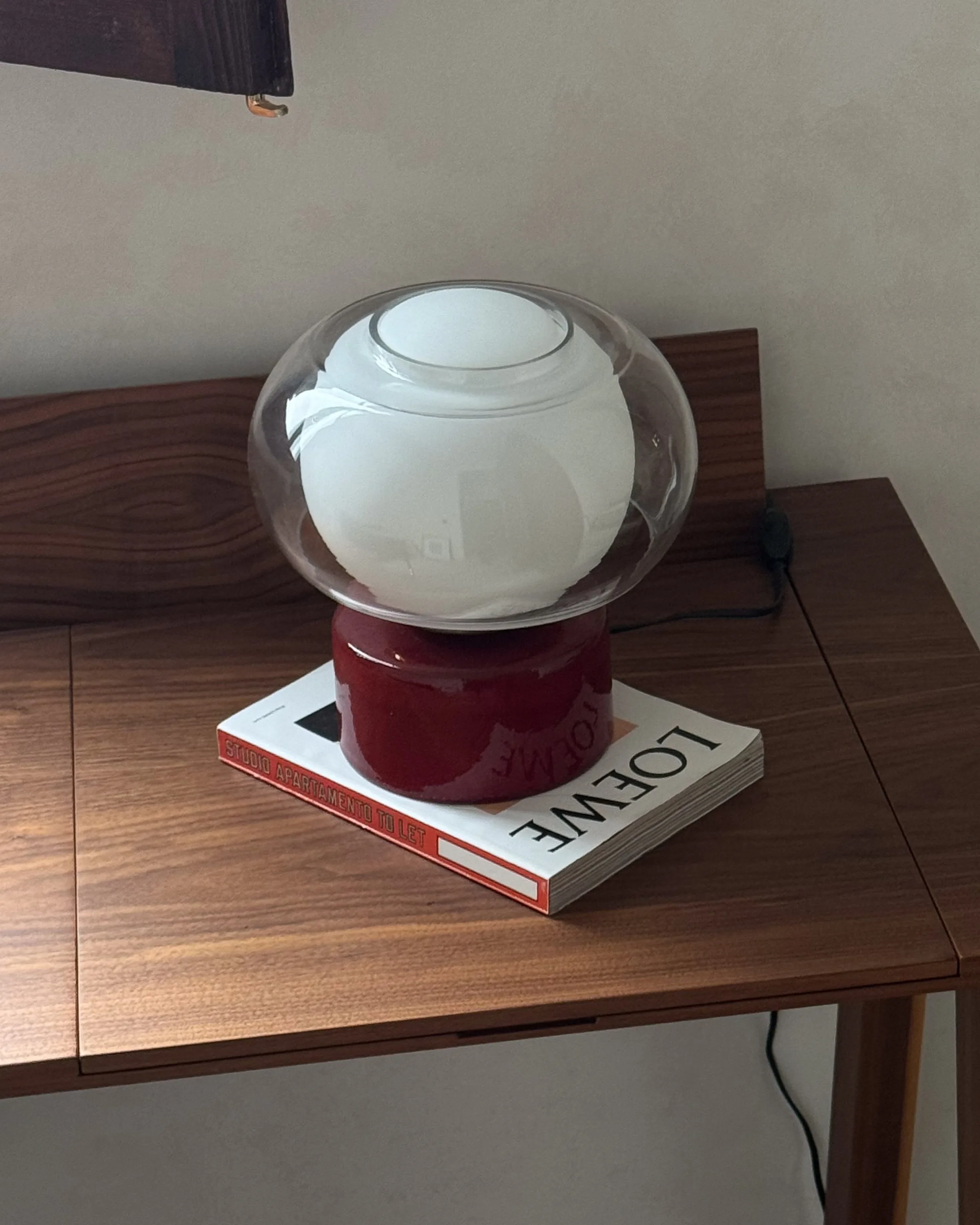 Table lamp with a glossy red base and clear glass globe on a Loewe book atop a walnut desk.