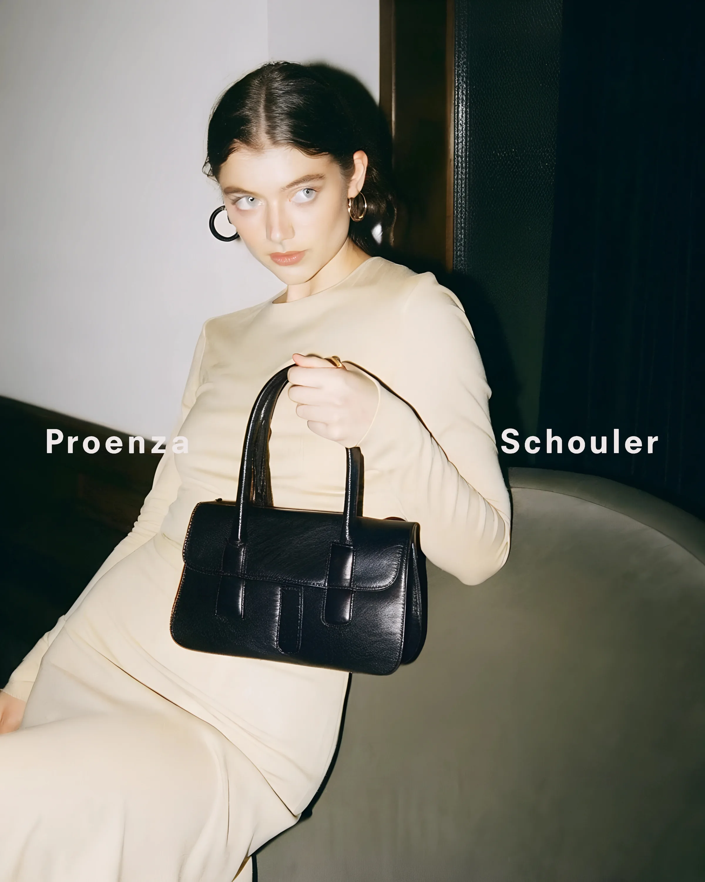 AI image of a model in a cream knit dress holding a black leather top-handle bag on a velvet sofa. Proenza Schouler AI Concept.
