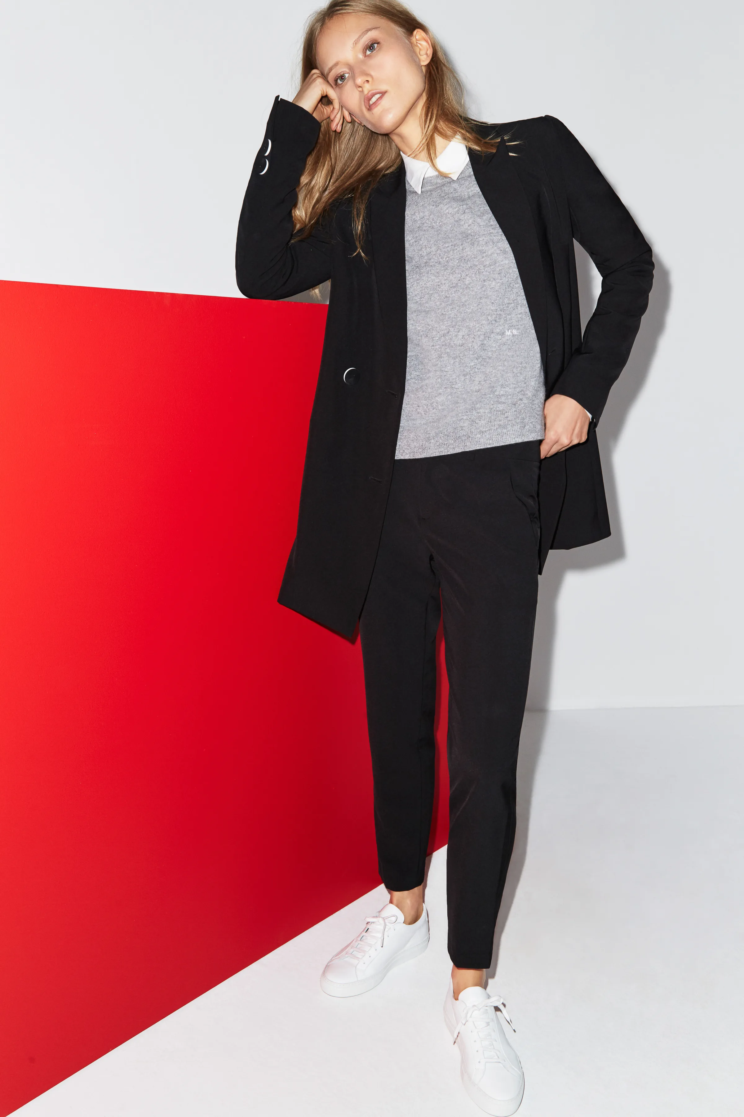 Model in a long black blazer, grey sweater and white sneakers leaning on a red panel in a white studio, for Misha Nonoo.