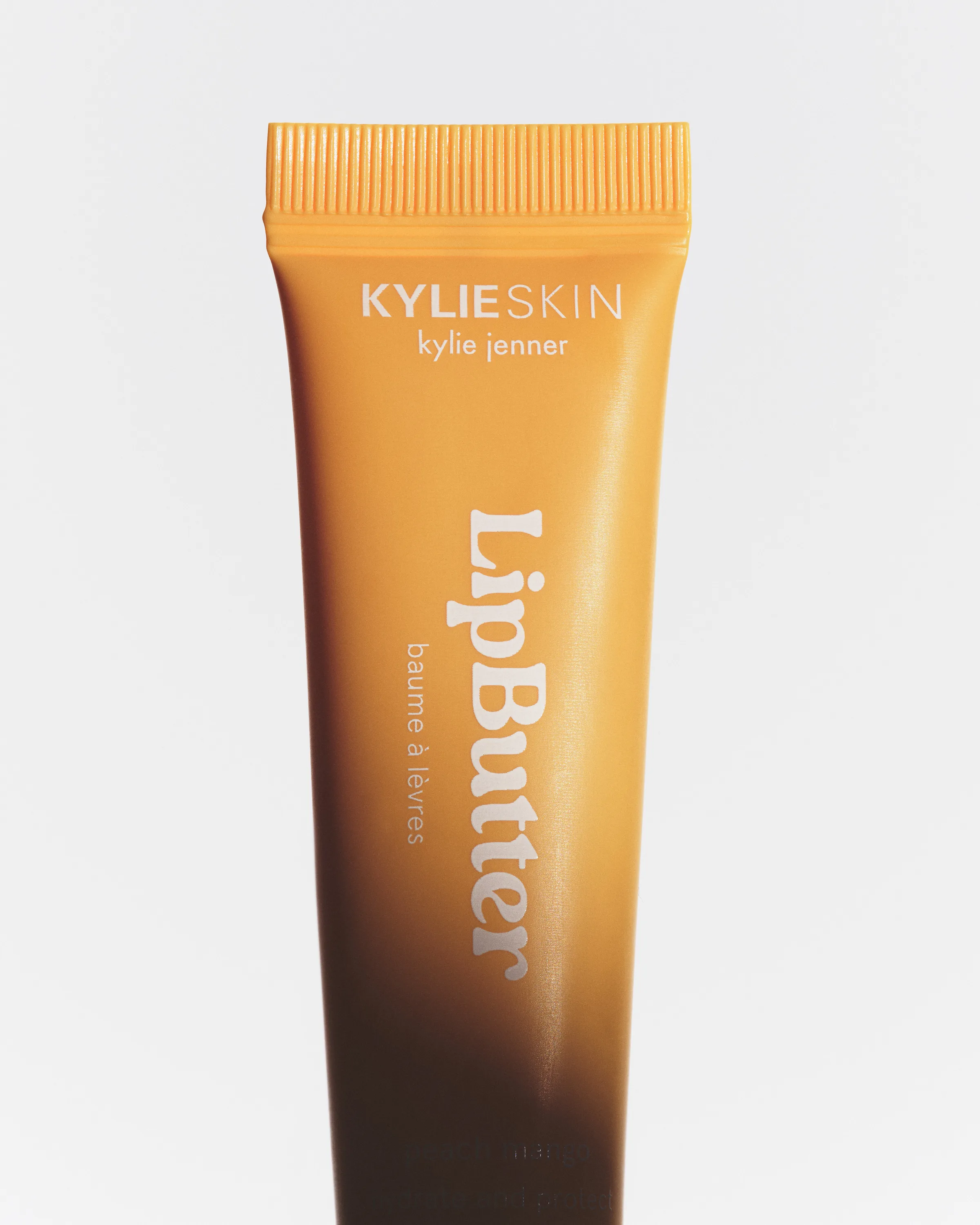 Close-up of an orange-to-brown gradient Lip Butter tube on a pale grey background, for Kylie Skin.