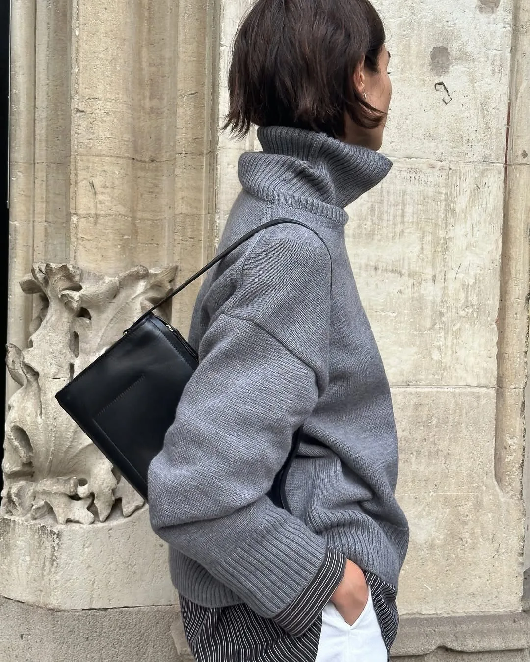 Woman in an oversized grey turtleneck sweater with a black leather shoulder bag beside a carved stone wall.
