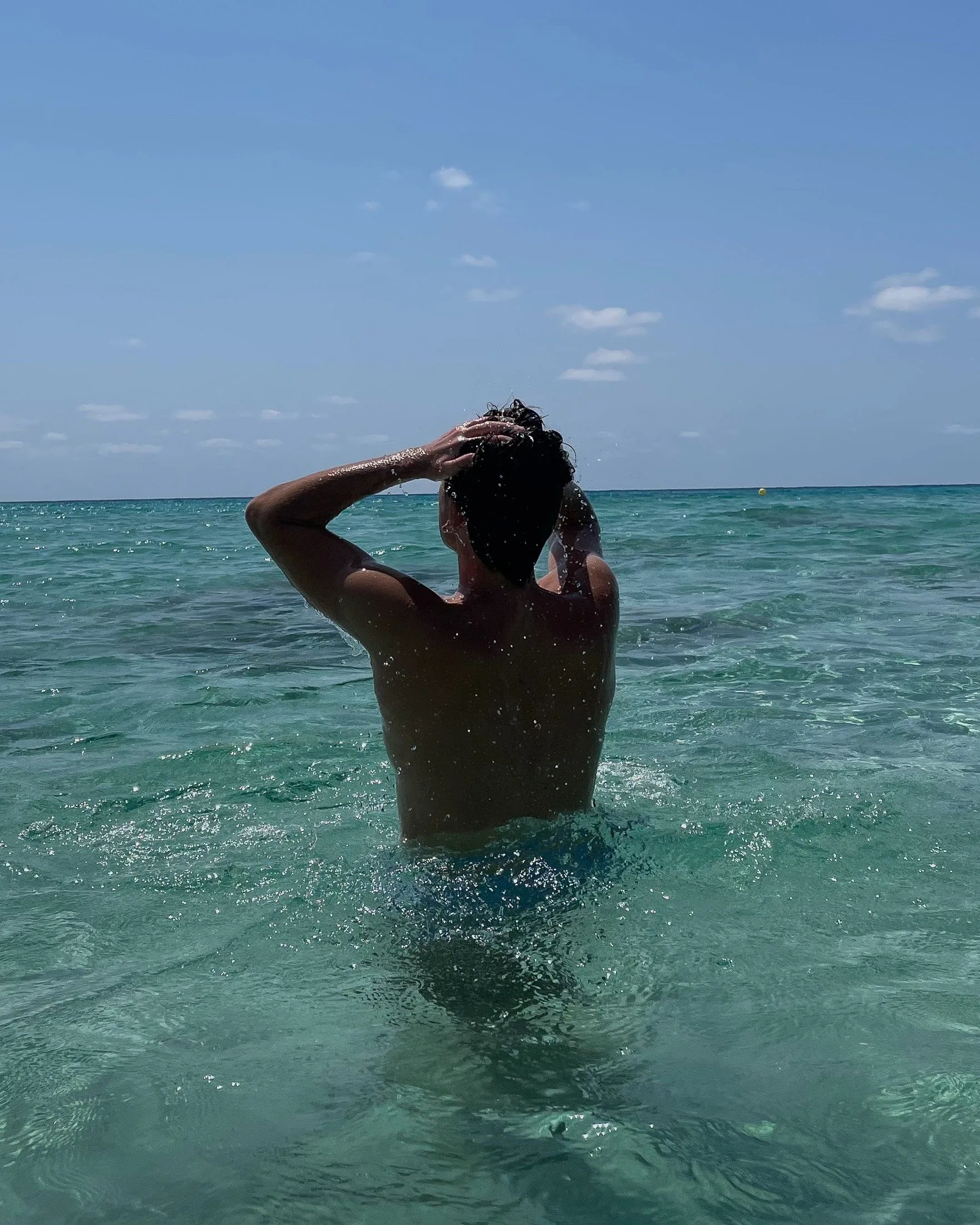 Man seen from behind waist-deep in clear turquoise sea, hands in wet hair under a blue sky.