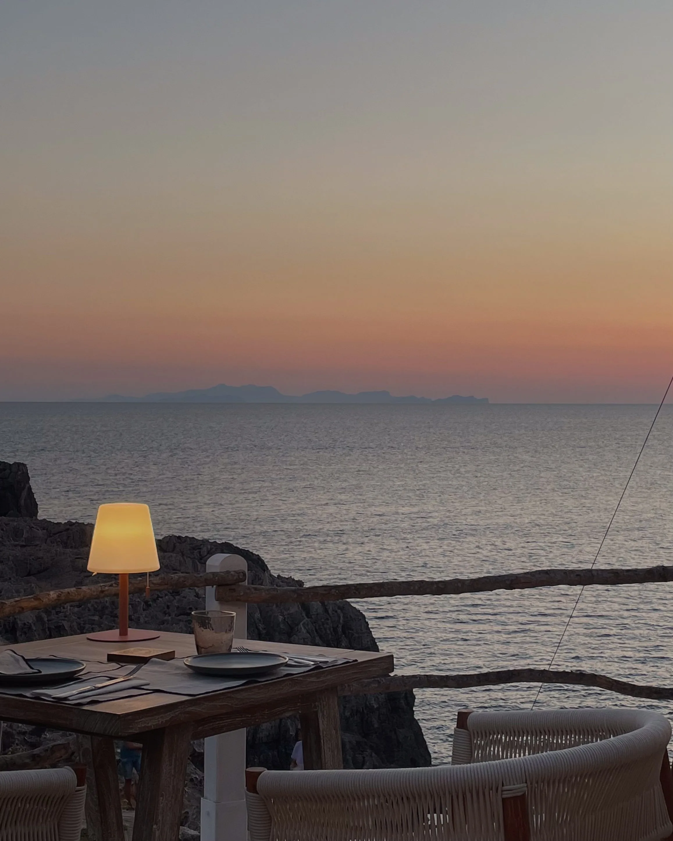 Outdoor restaurant table with glowing lamp on a cliff overlooking the sea at sunset, wicker chairs in front.