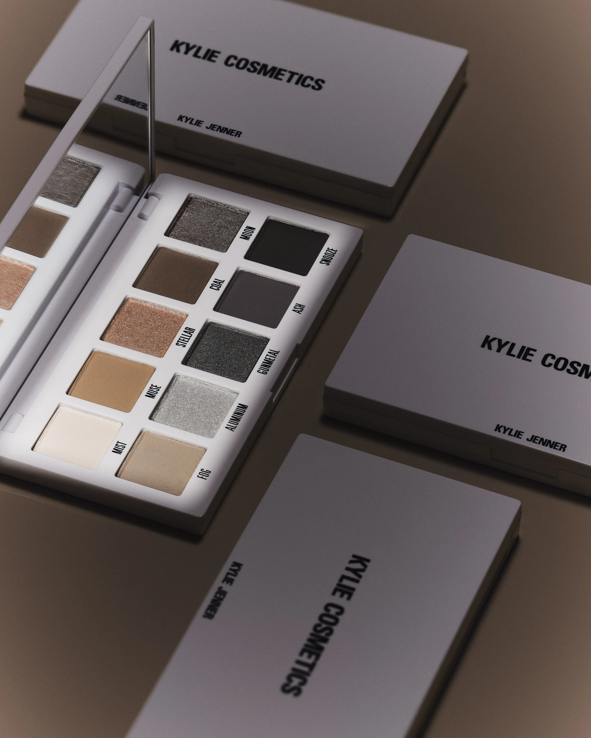 Open smoky eyeshadow palette in grey, taupe and silver shades beside closed grey compacts on a brown backdrop, for Kylie Cosmetics.