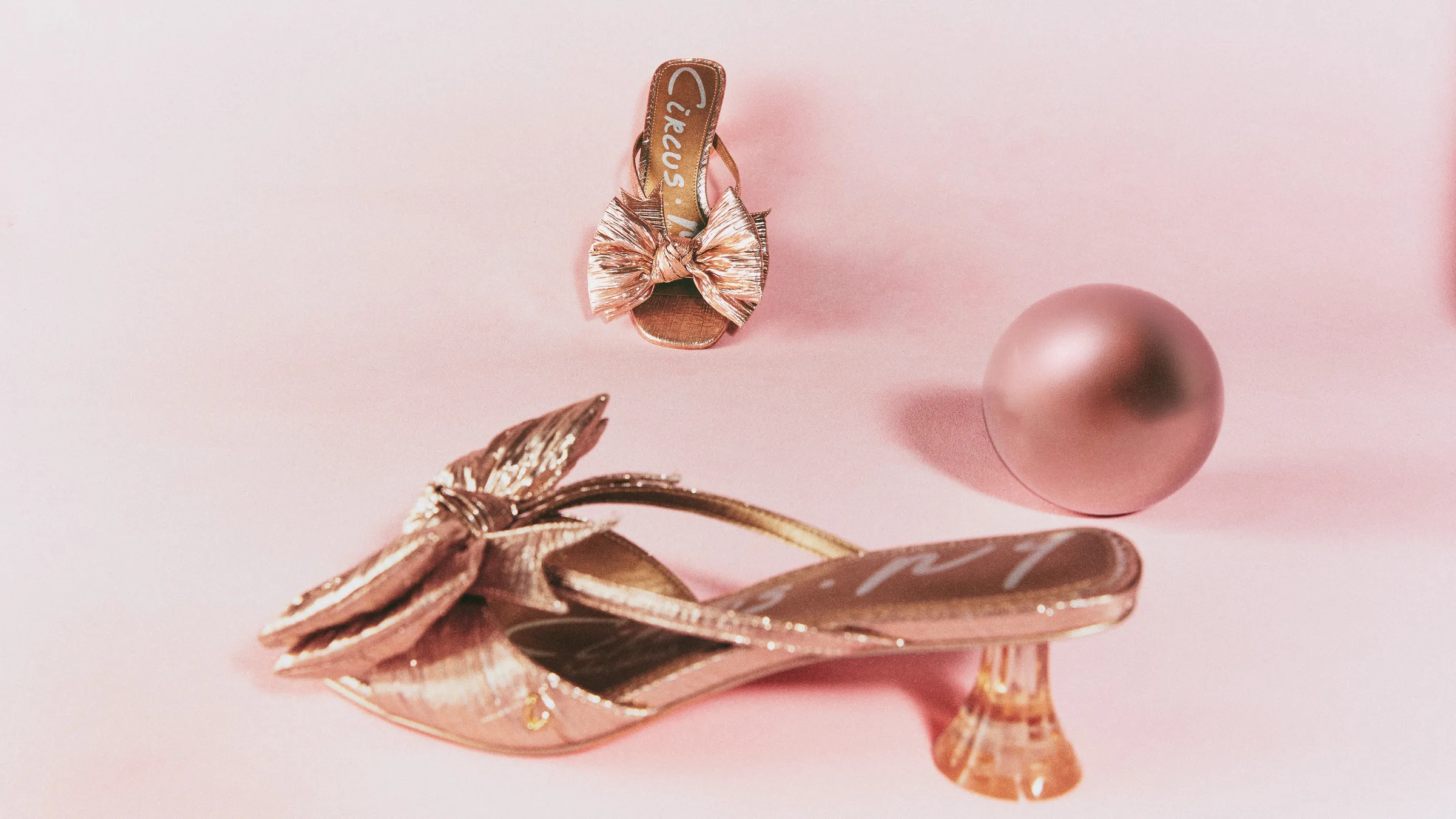 Metallic gold bow mules with a lucite heel beside a rose-gold sphere on a pink backdrop, for Circus NY.