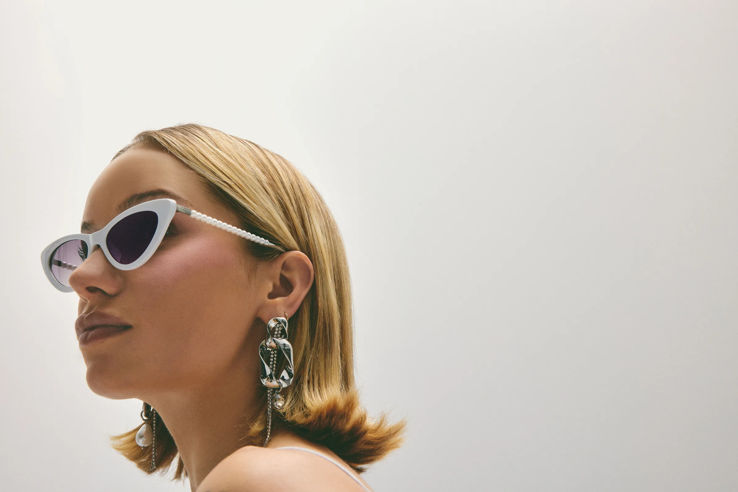 Blonde model in profile wearing white cat-eye sunglasses with pearl arms and silver chain earrings, for Sam Edelman.