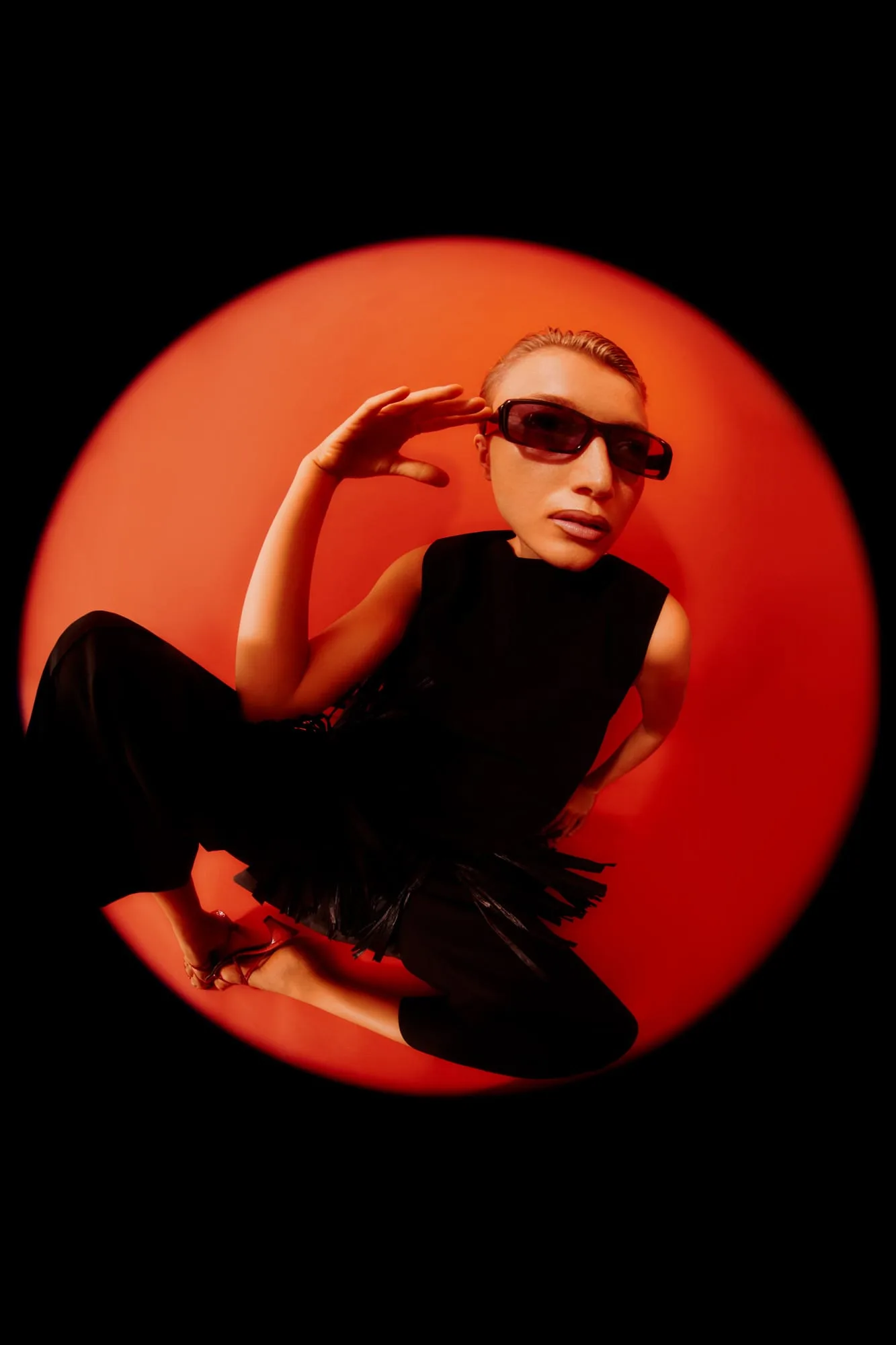 Model in black sleeveless dress and dark sunglasses crouching in a red circular spotlight, fisheye view, for Numéro.