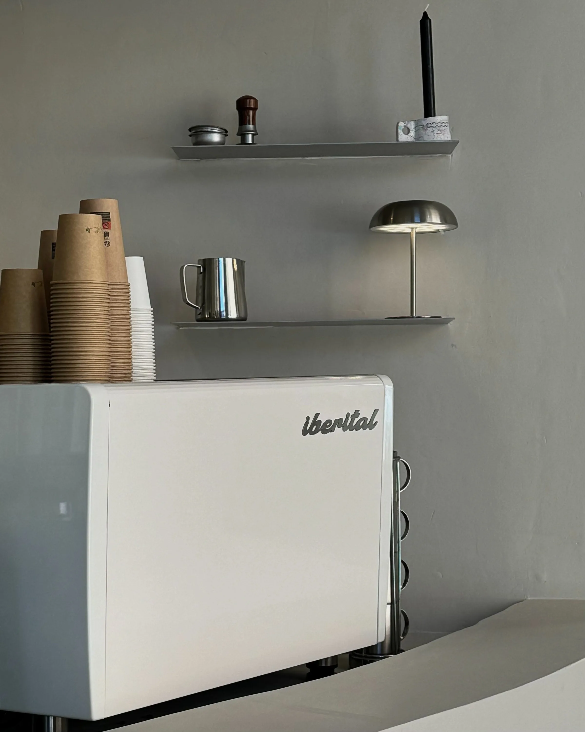 White Iberital espresso machine with stacked kraft cups, steel shelves holding a milk jug, lamp and candle.