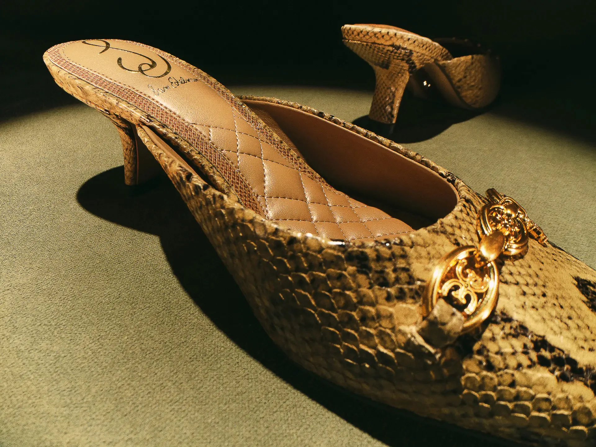 Snakeskin kitten-heel mules with gold logo hardware and quilted insole on green felt under dramatic light, for Sam Edelman.