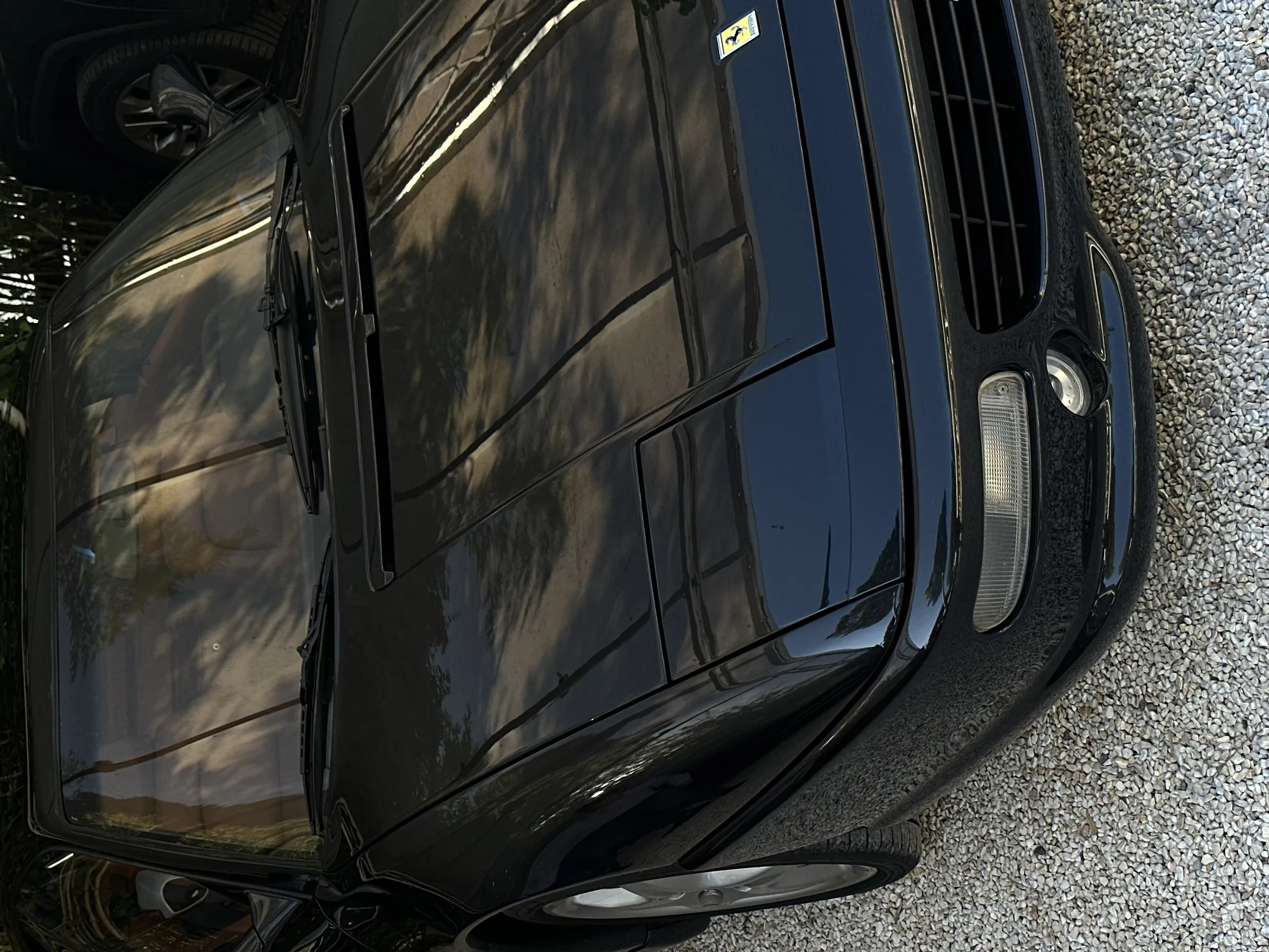 Black Ferrari sports car parked on gravel, dappled shade from trees reflecting across its glossy hood.