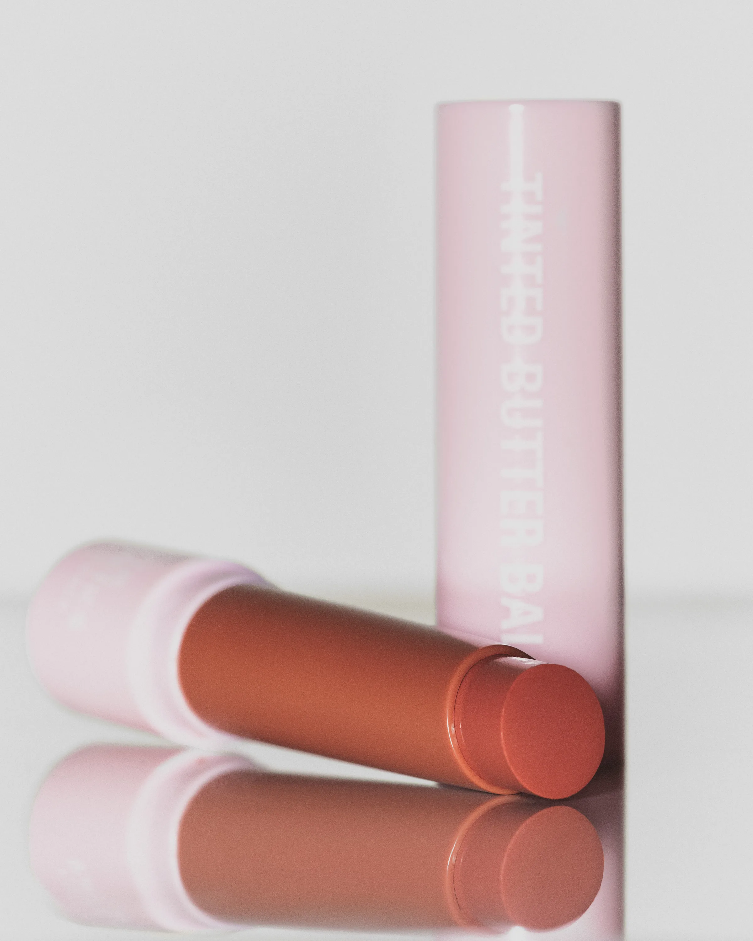 Tinted butter balm in a honey shade lying beside its pink cap on a reflective white surface, for Kylie Cosmetics.