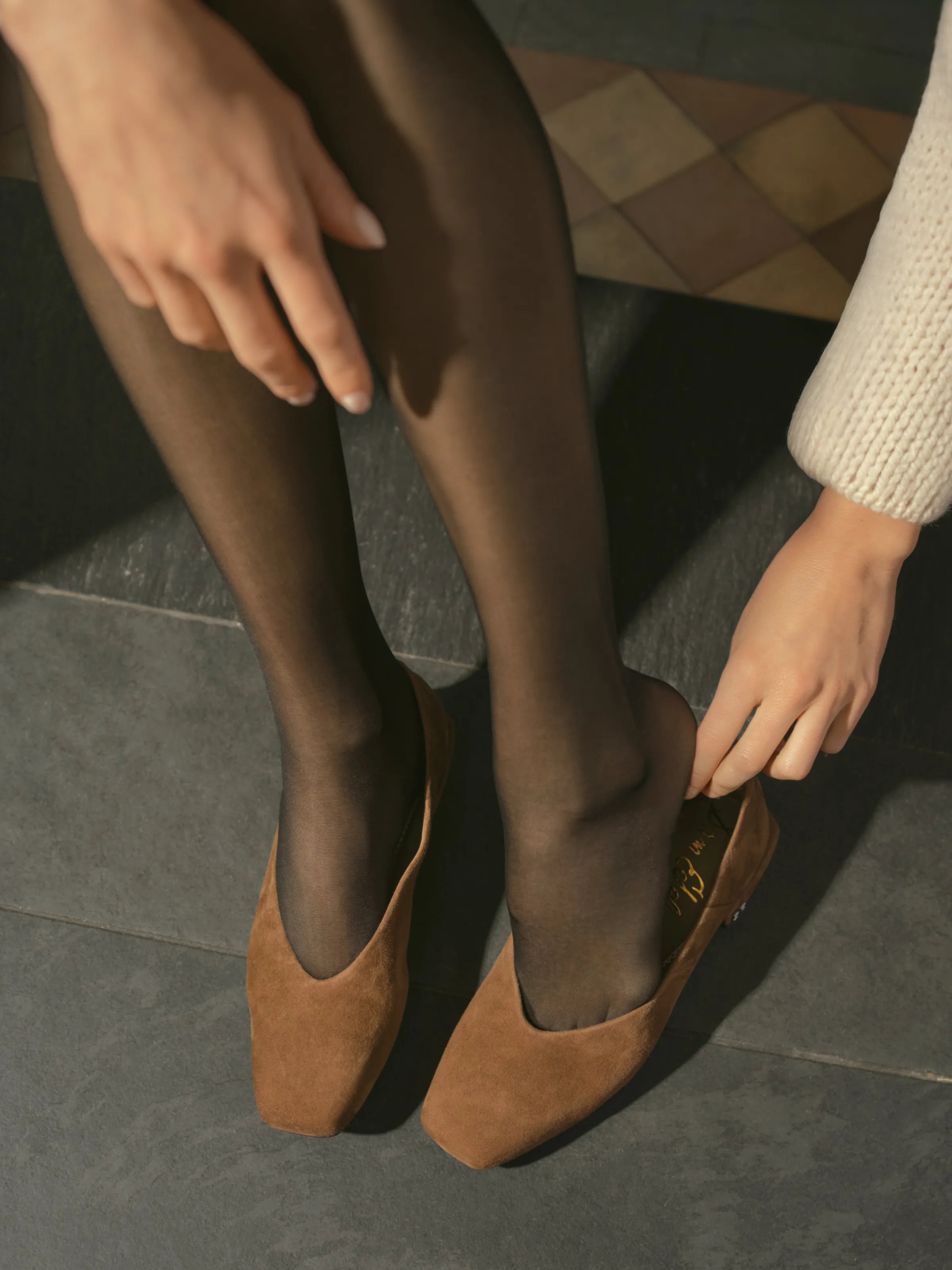 Model in sheer black tights slipping on tan suede square-toe flats on a grey stone floor, for Sam Edelman.
