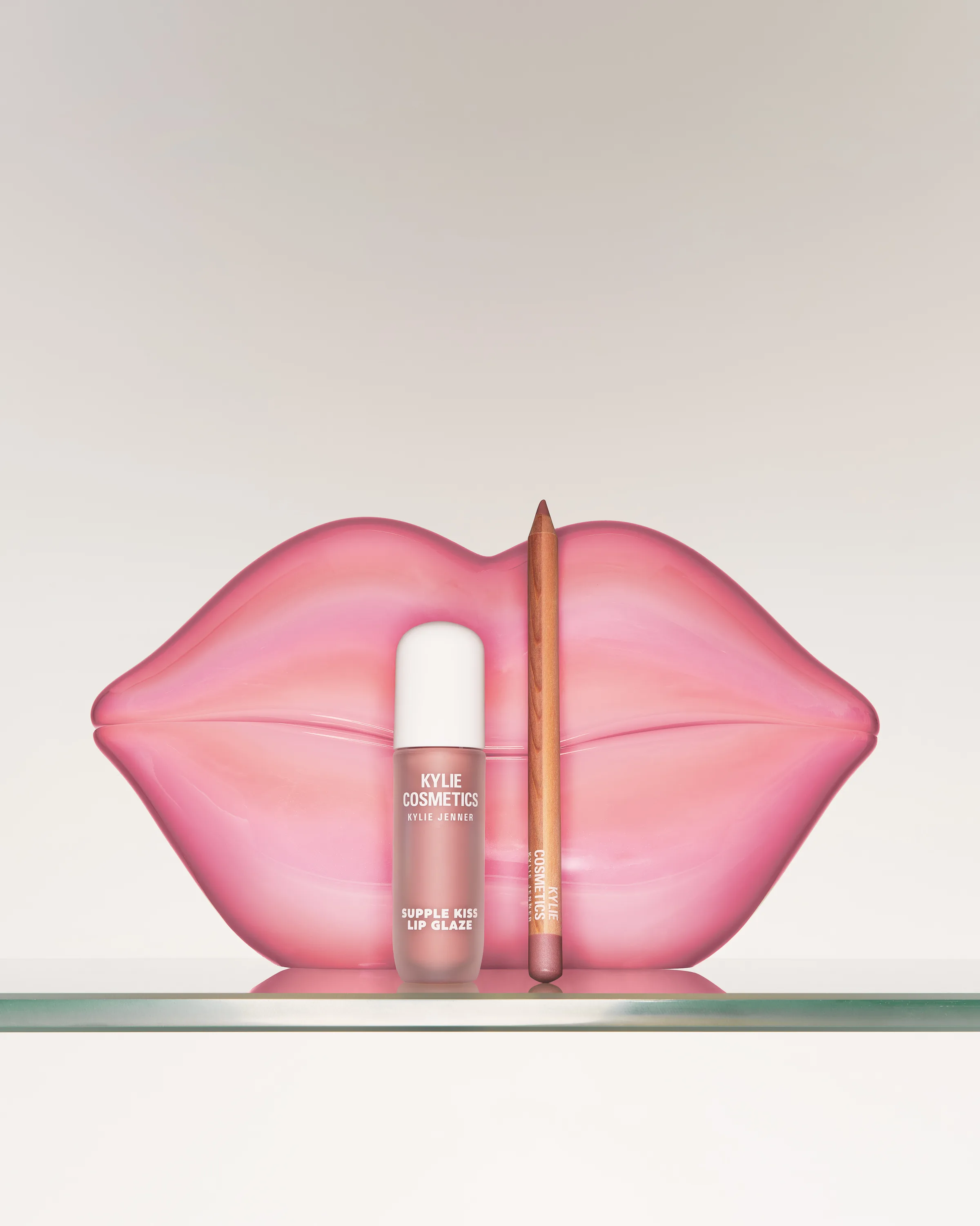 Supple Kiss Lip Glaze and lip liner in front of a glossy pink lips sculpture on a glass shelf, for Kylie Cosmetics.