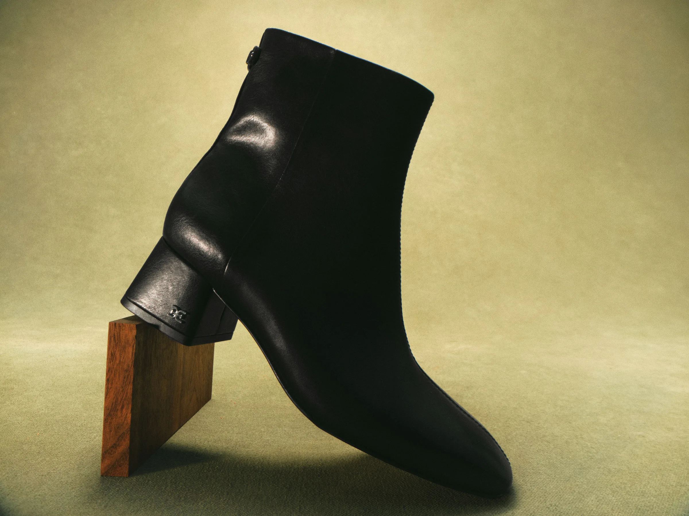 Black leather ankle boot with block heel propped on a wooden block against a sage green backdrop, for Sam Edelman.
