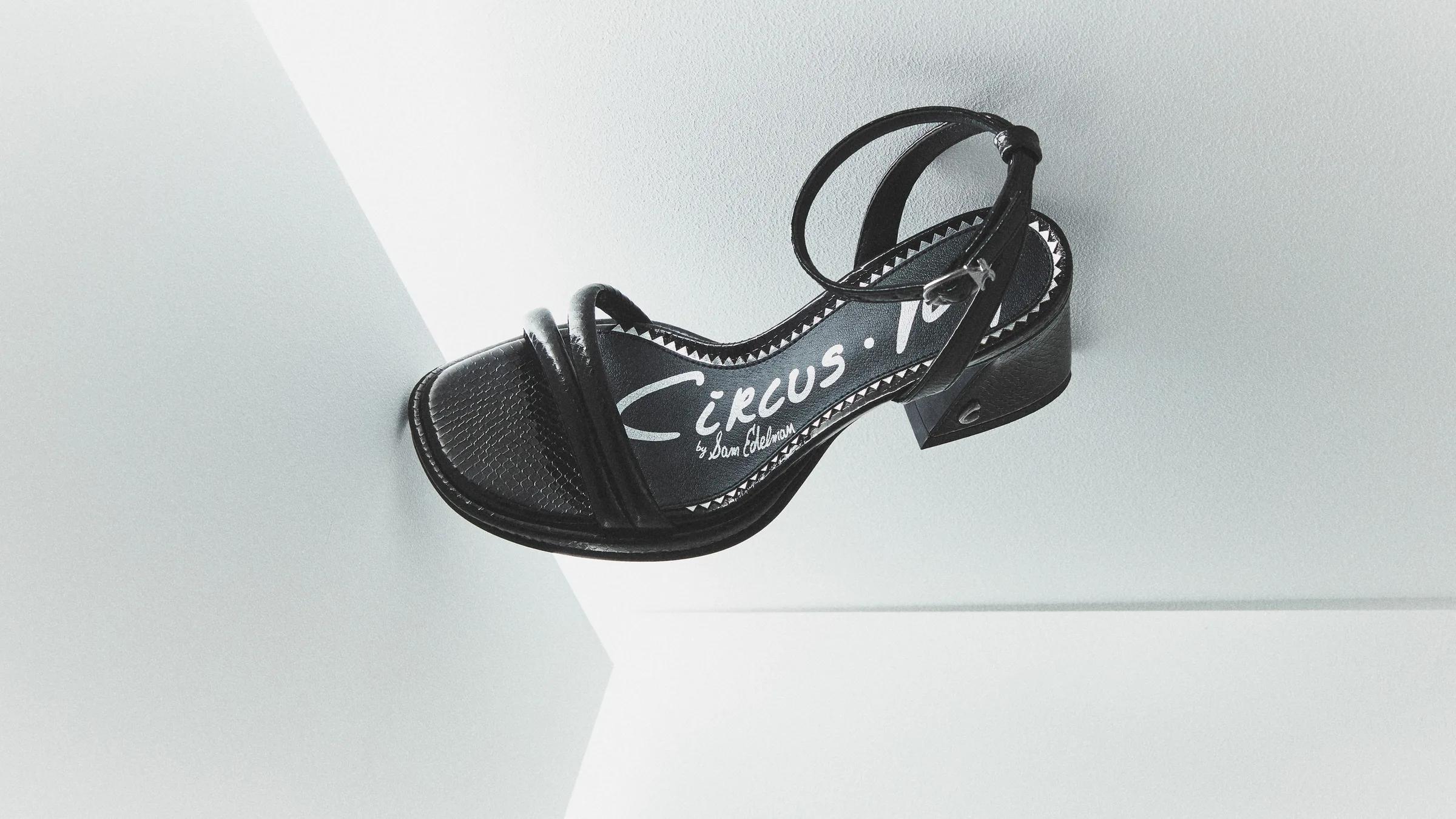 Black strappy block-heel sandal with ankle strap, angled against white geometric walls, for Circus NY.
