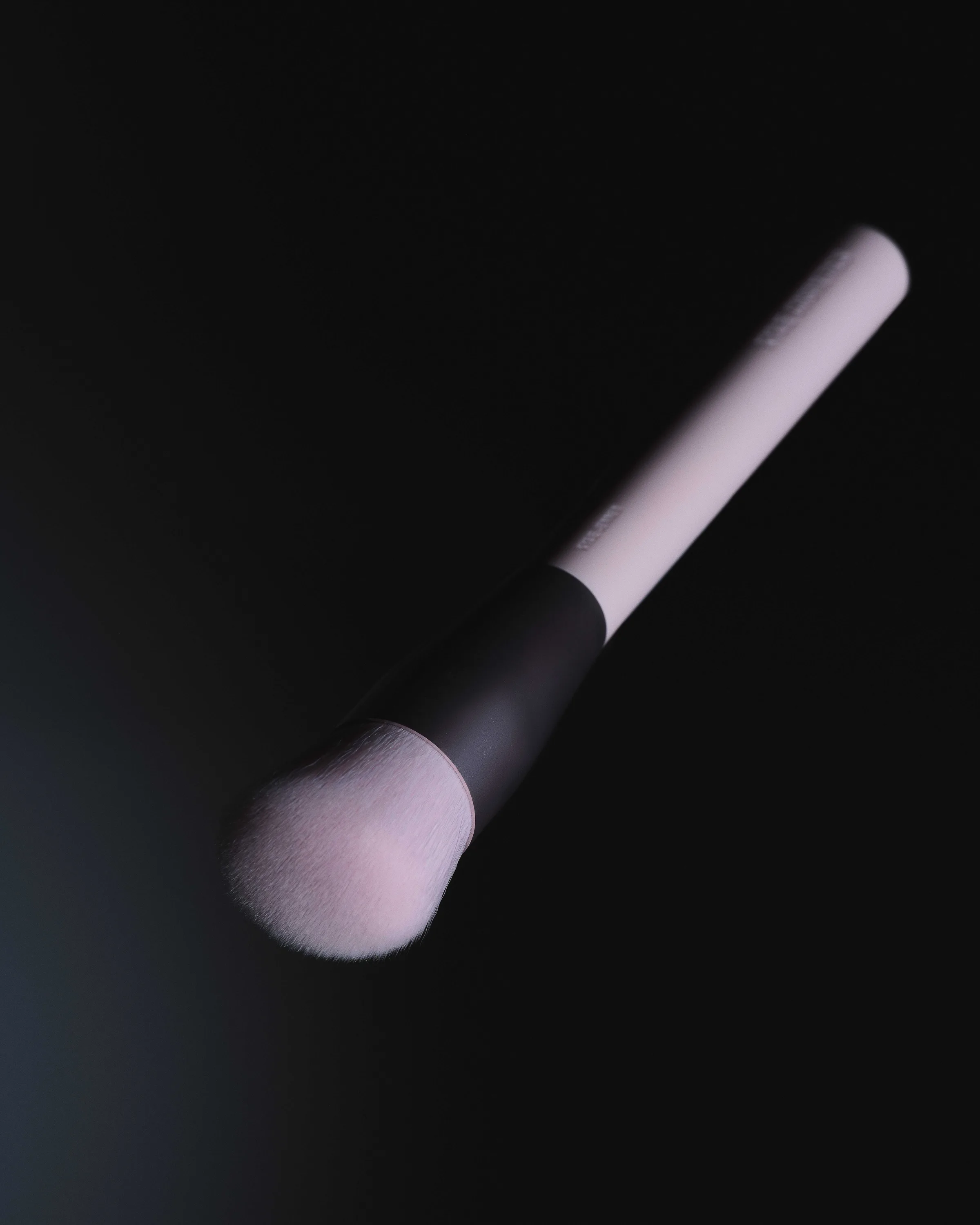 Pale pink makeup brush with a black ferrule floating diagonally against a dark background, for Kylie Cosmetics.