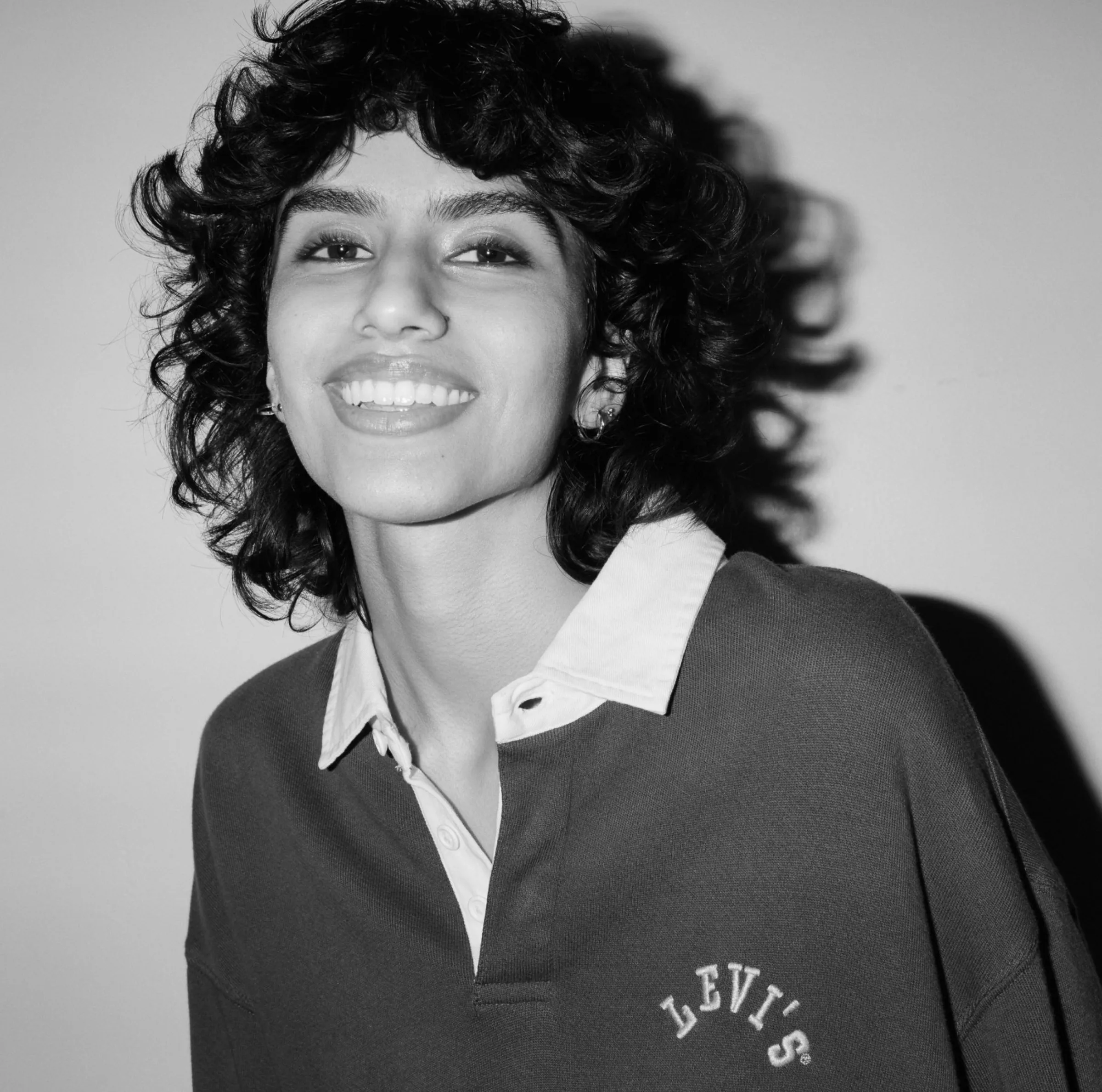 Black and white portrait of a smiling curly-haired model in a collared rugby sweatshirt with embroidered logo, for Levi's.