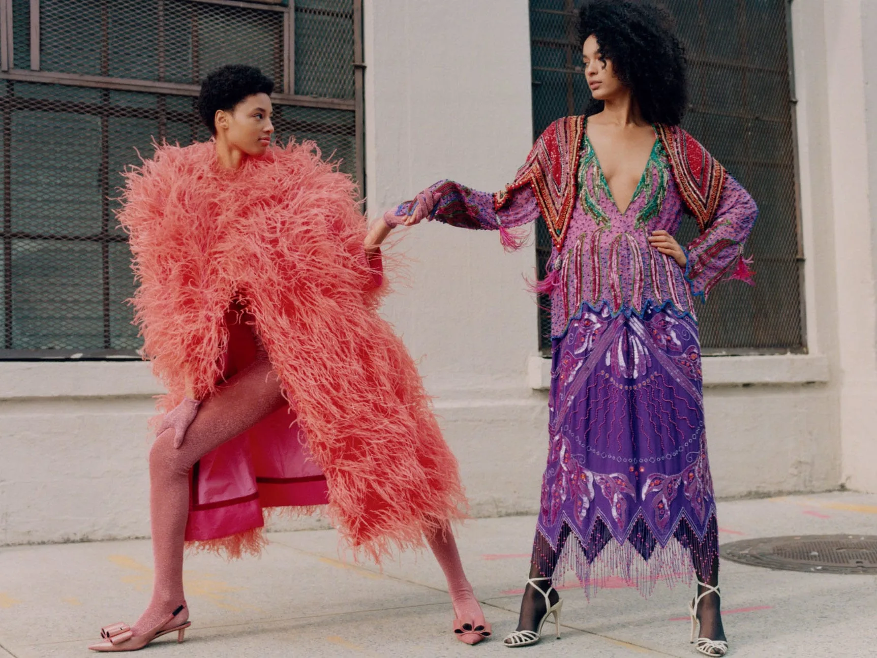 Two models on a city sidewalk holding hands, one in a coral feather coat, the other in a purple beaded gown, for Vogue Mexico.