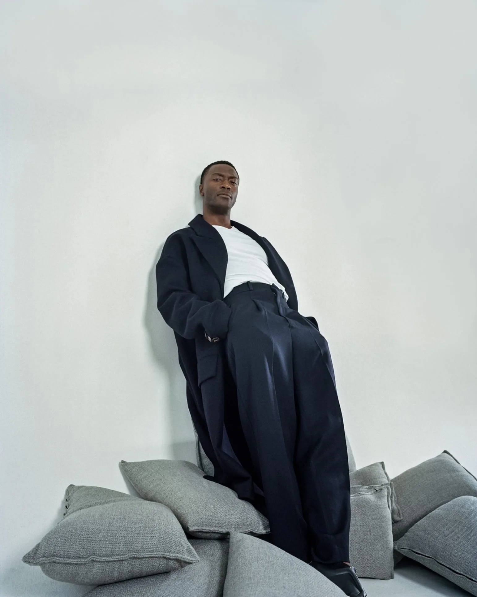 Man in a navy overcoat, wide trousers and white tee leaning against a white wall on grey cushions, for The Last Magazine.