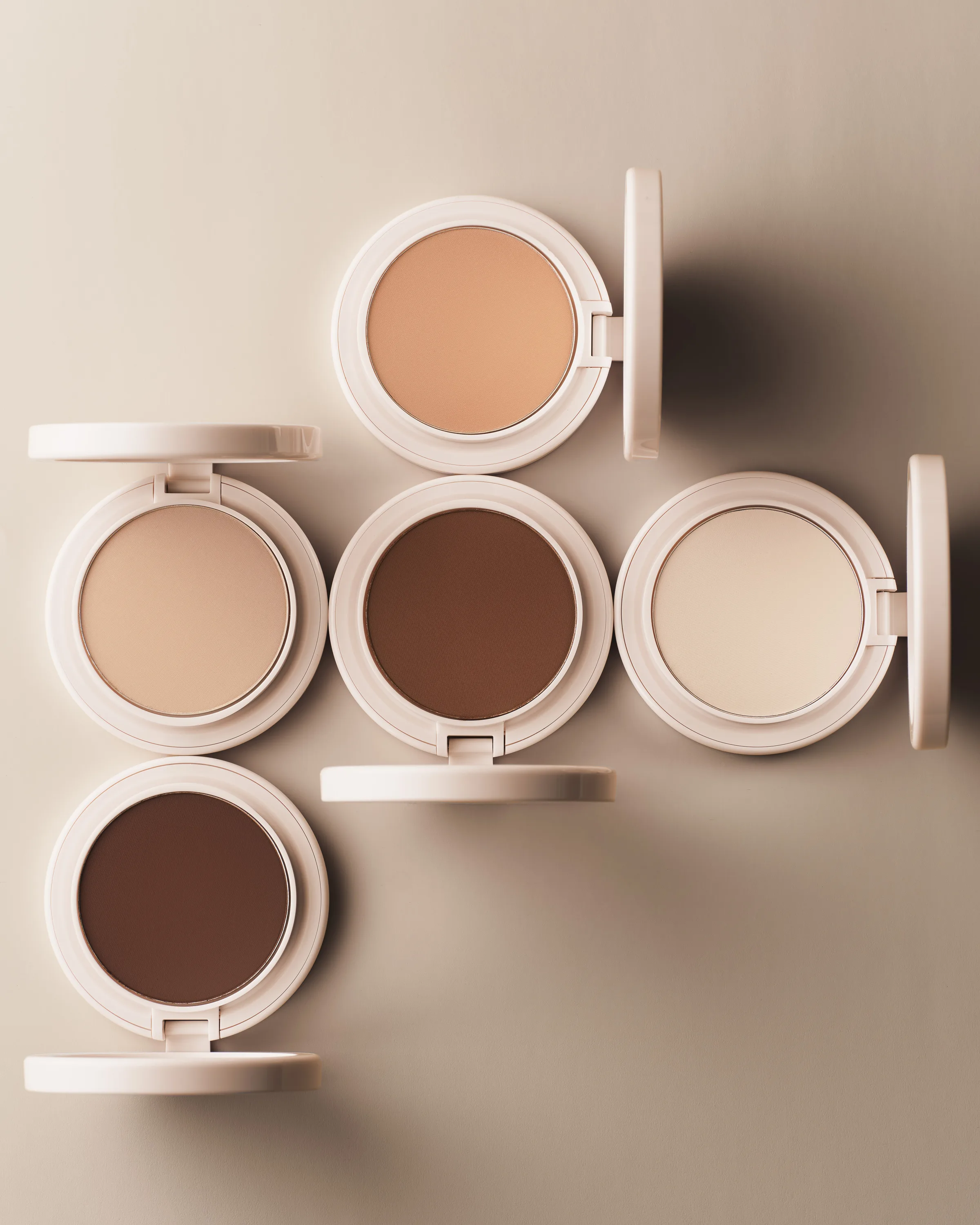 Five open cream powder compacts in shades from ivory to deep brown, arranged on a beige surface, for Kylie Cosmetics.