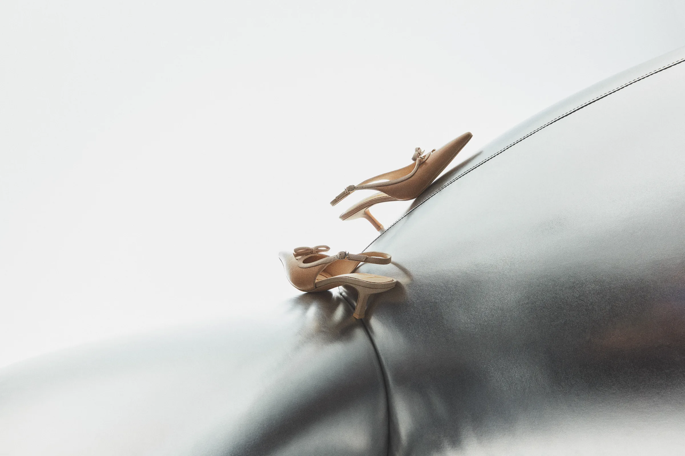 Beige bow-trimmed slingback kitten heels perched on a curved chrome surface against white, for Sam Edelman.