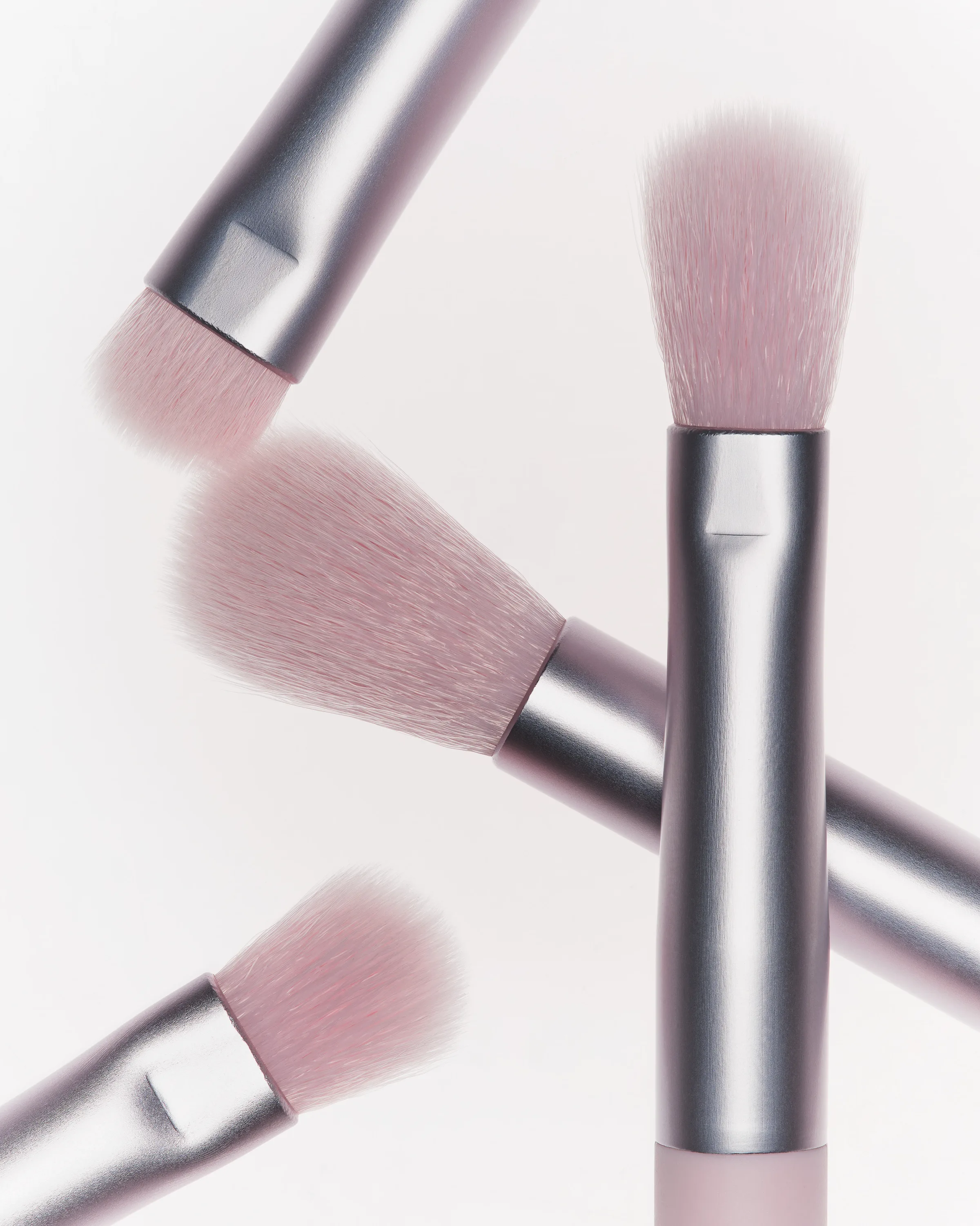 Four pink-bristled makeup brushes with satin silver ferrules crossing on a white background, for Kylie Cosmetics.