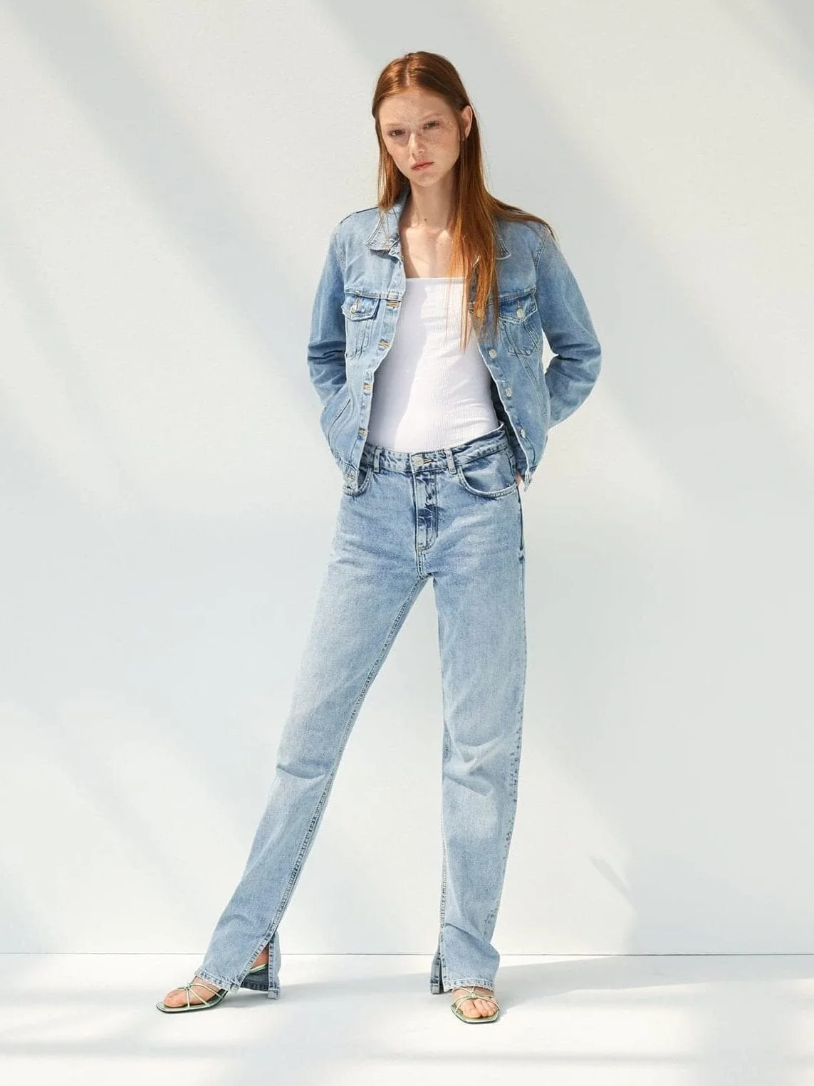 Red-haired model in a light wash denim jacket, white tank and split-hem jeans against a sunlit white wall, for Zara.