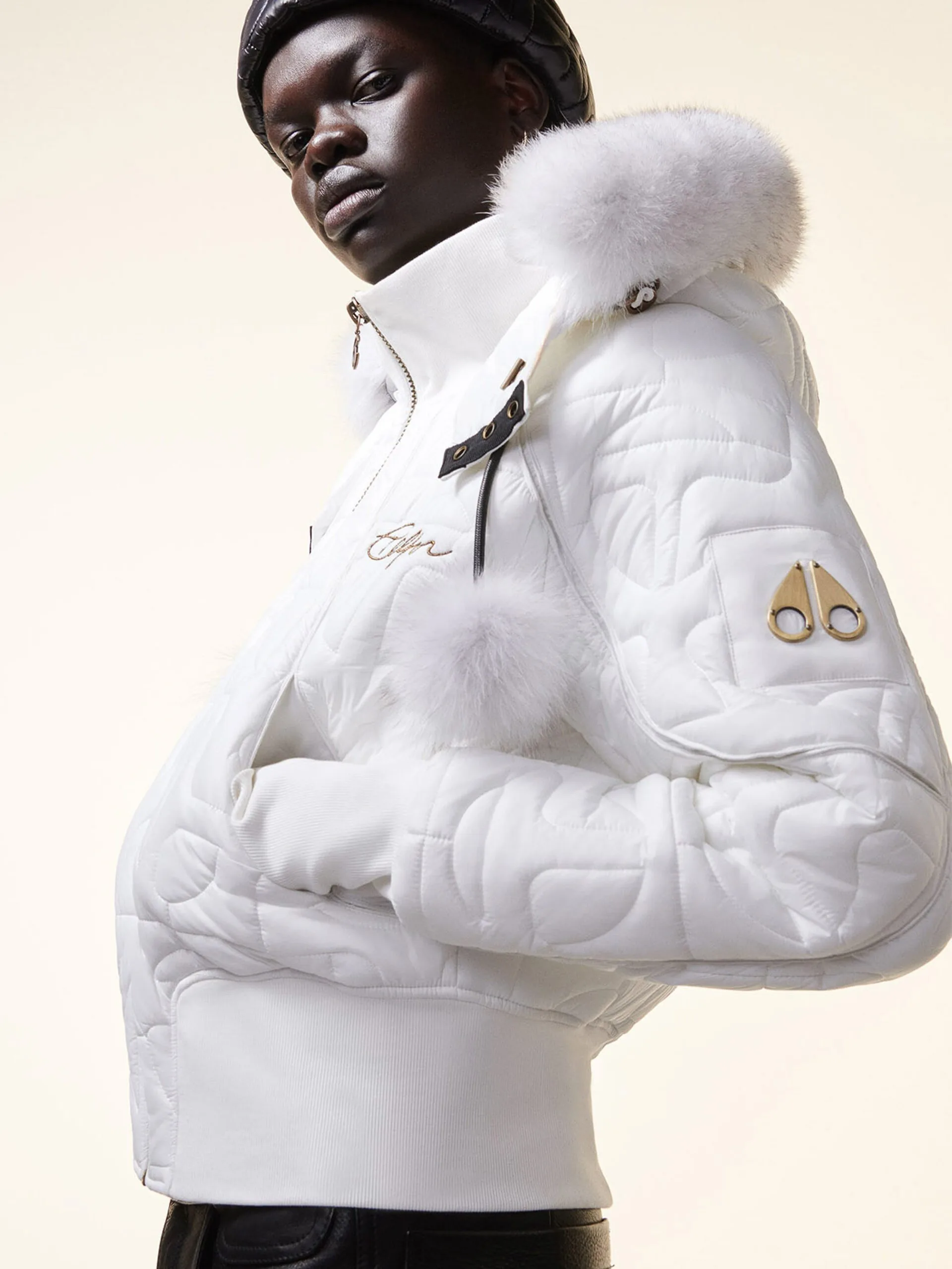 Model in a white quilted bomber jacket with fur-trimmed hood and pom-poms, gold logo on sleeve, for Moose Knuckles.
