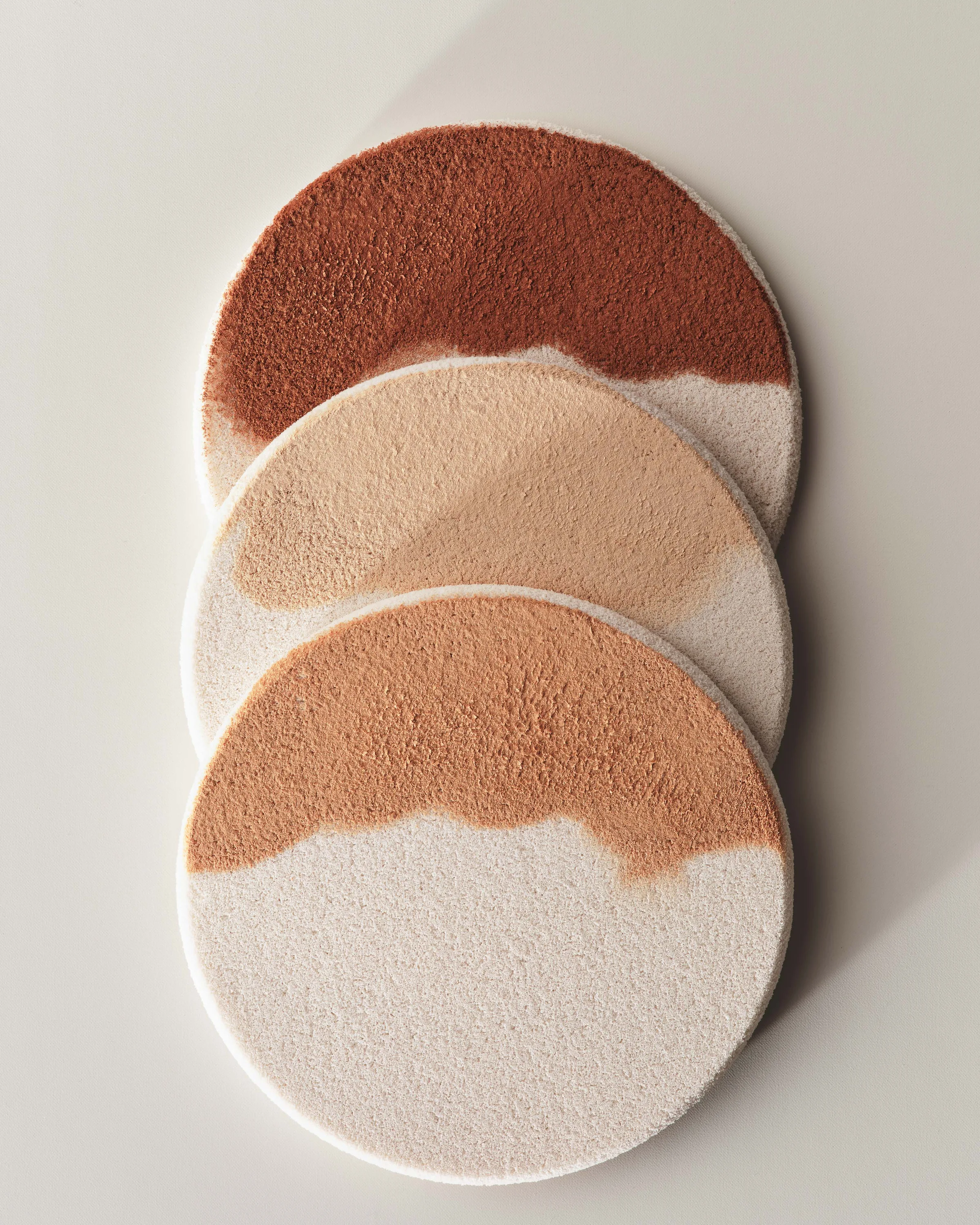 Three round makeup sponges overlapping, each half-coated in a different foundation shade, on a cream backdrop, for Kylie Cosmetics.