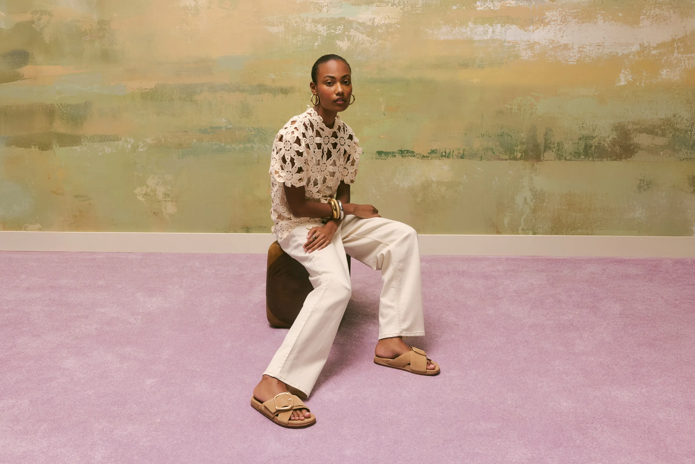 Model in a crochet top and white jeans sits on a stool wearing tan buckle slide sandals on a lilac carpet, for Sam Edelman.