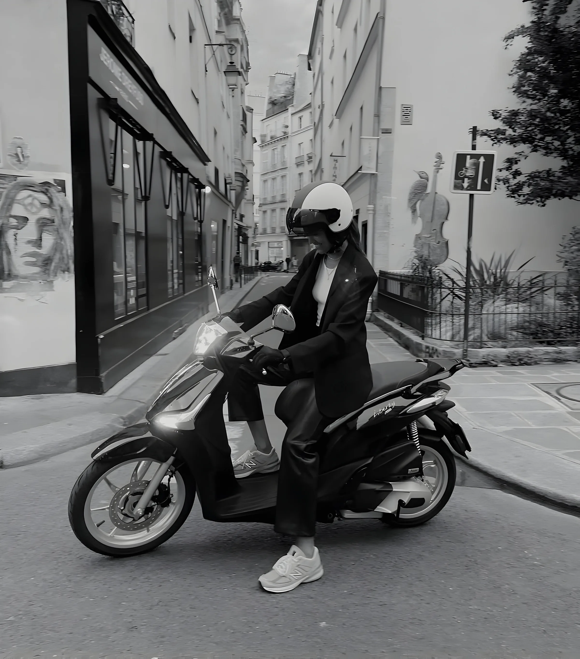 Black-and-white shot of a woman in a helmet, blazer and New Balance sneakers riding a scooter on a Paris street.