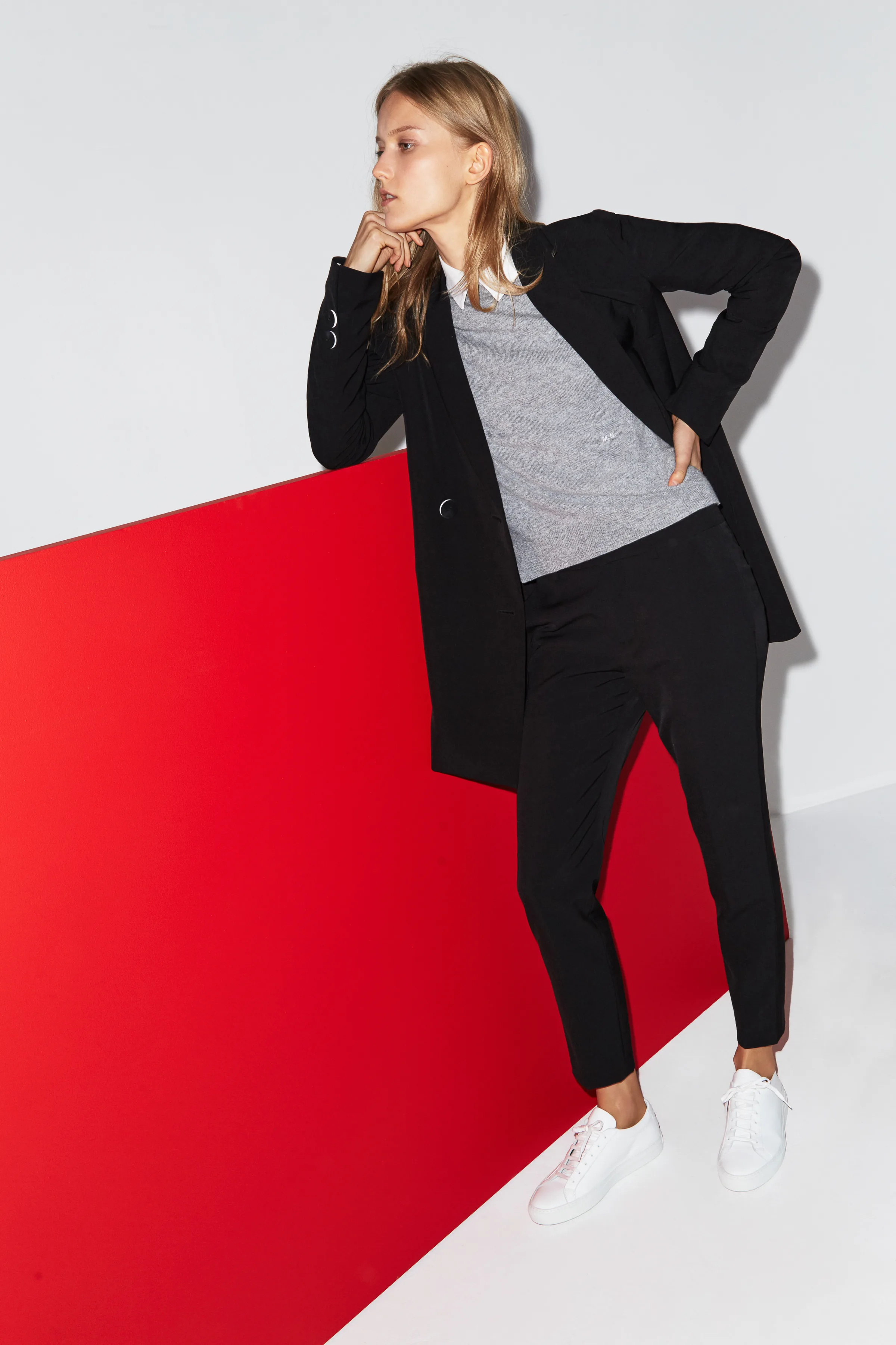 Model in black blazer, grey sweater, black trousers and white sneakers leaning on a red panel, for Misha Nonoo.