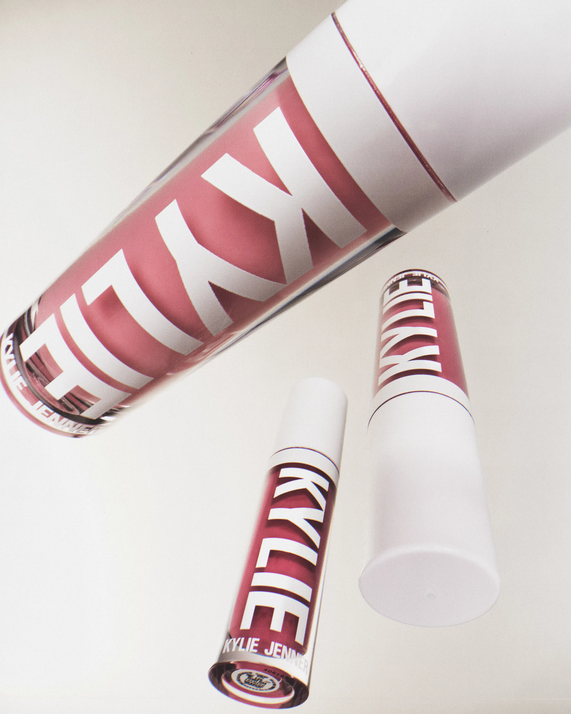 Three pink plumping lip gloss tubes with white caps floating against a white background, for Kylie Cosmetics.